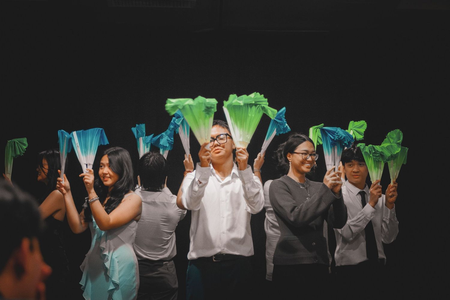 A group of performers smile and wave colorful blue and green fans on a dark stage.