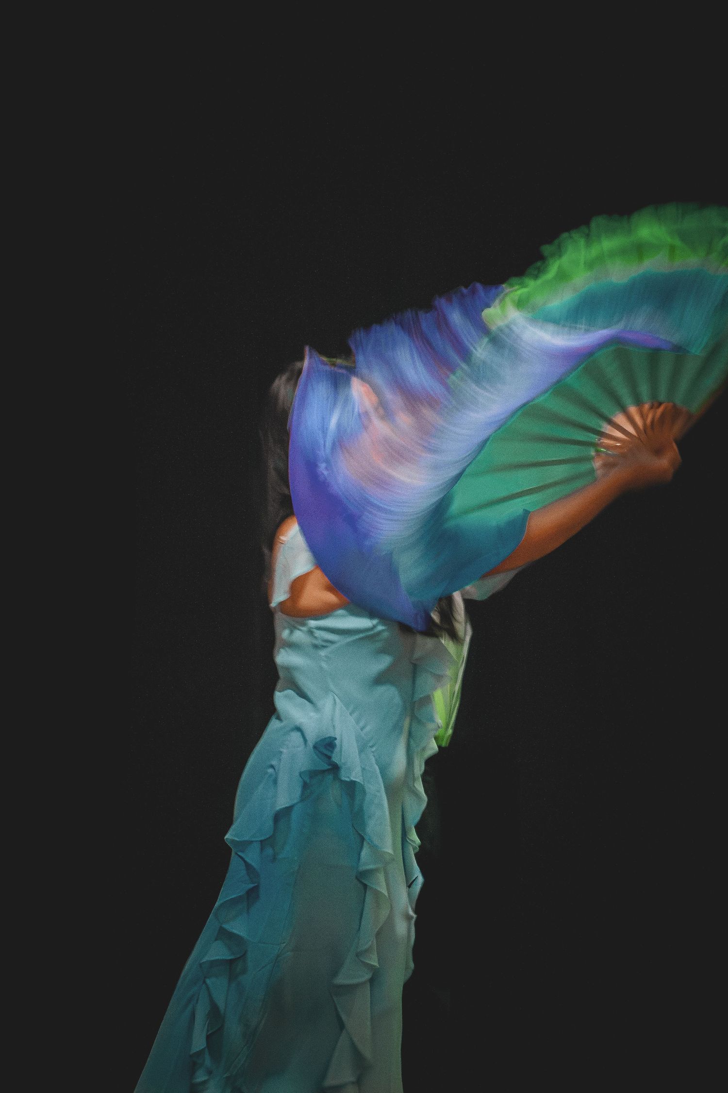 Back view of dancer in teal ruffled dress waving colorful blue and green silk fans.