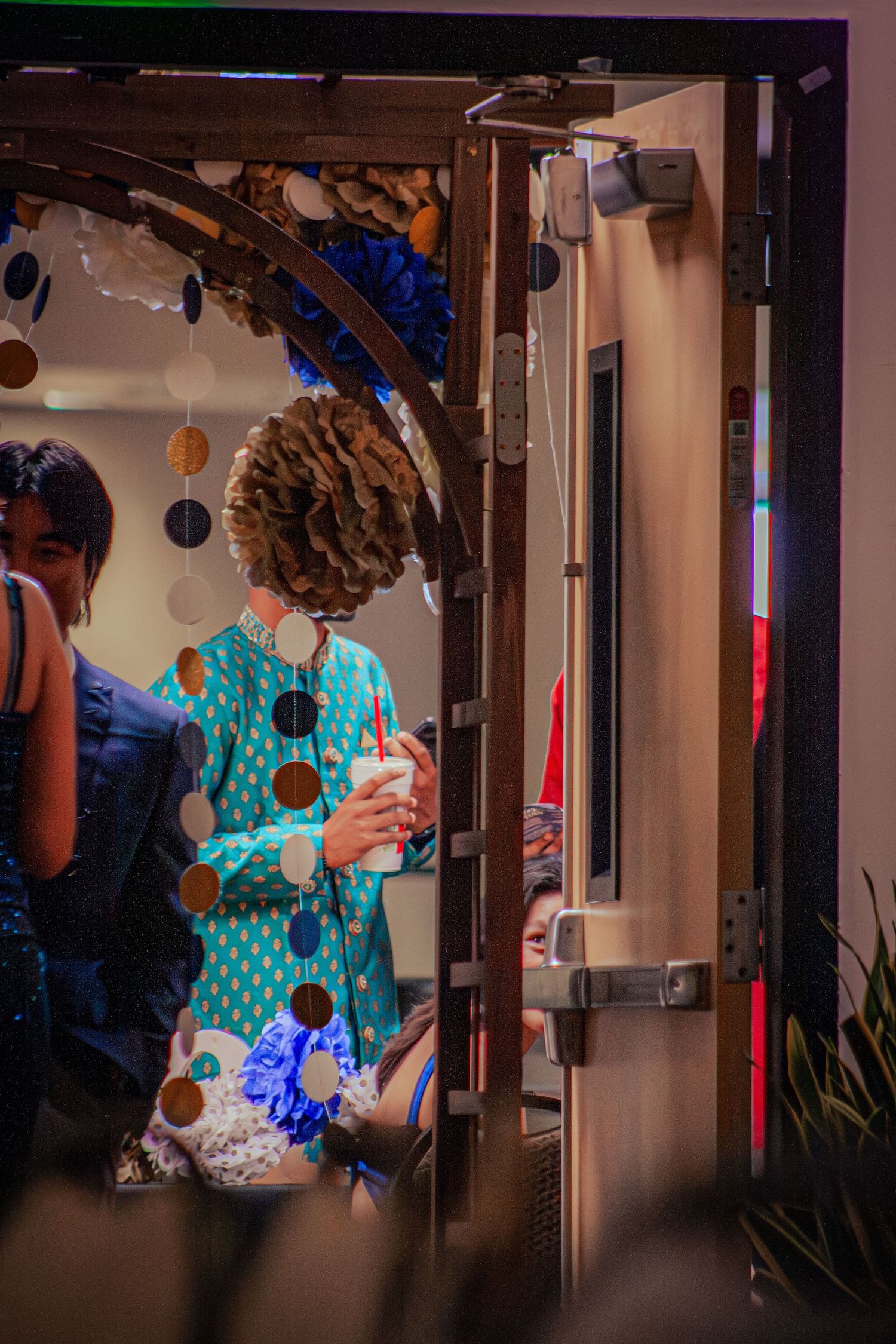 A person in a teal polka-dot outfit holds a drink near a decorated doorway with paper pompoms.