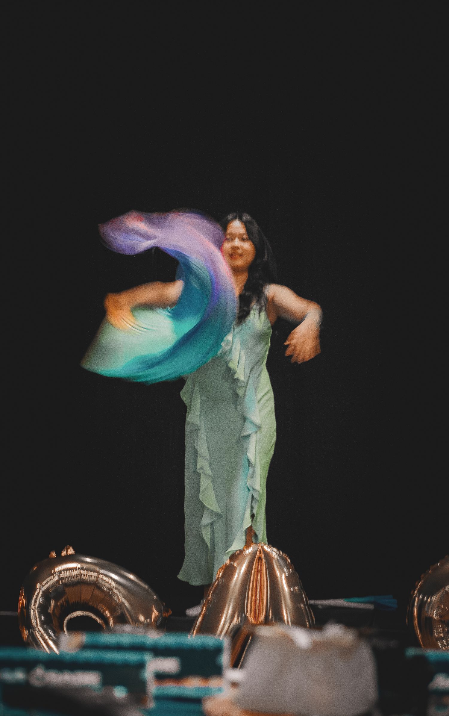 Young Asian woman in mint green ruffled dress performing dynamic silk fan dance on stage.