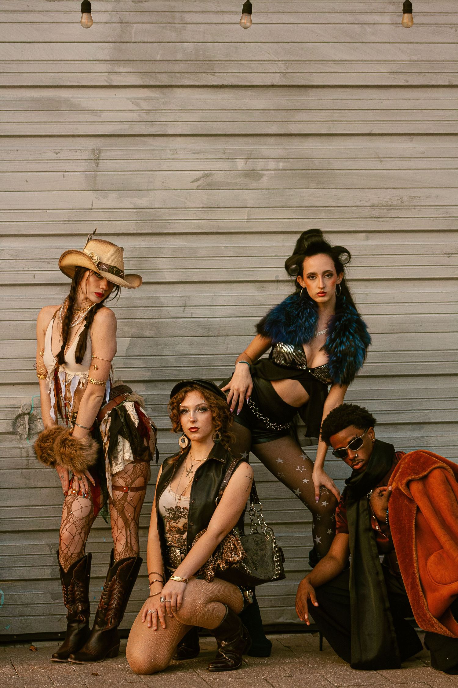 Four women in edgy post-apocalyptic costumes pose against a corrugated metal wall outdoors.