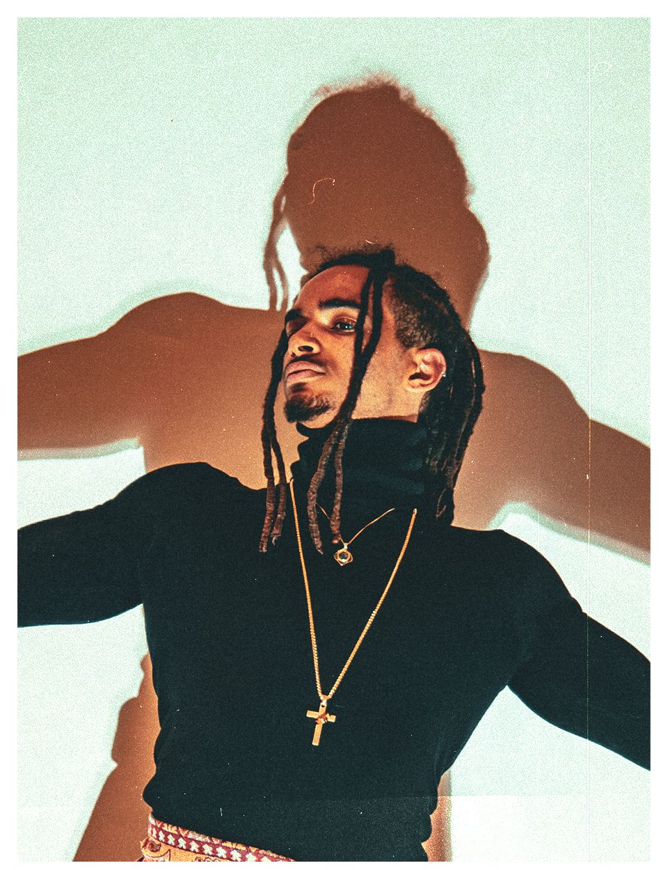 A man with dreadlocks poses dynamically wearing a black turtleneck and gold cross necklace.