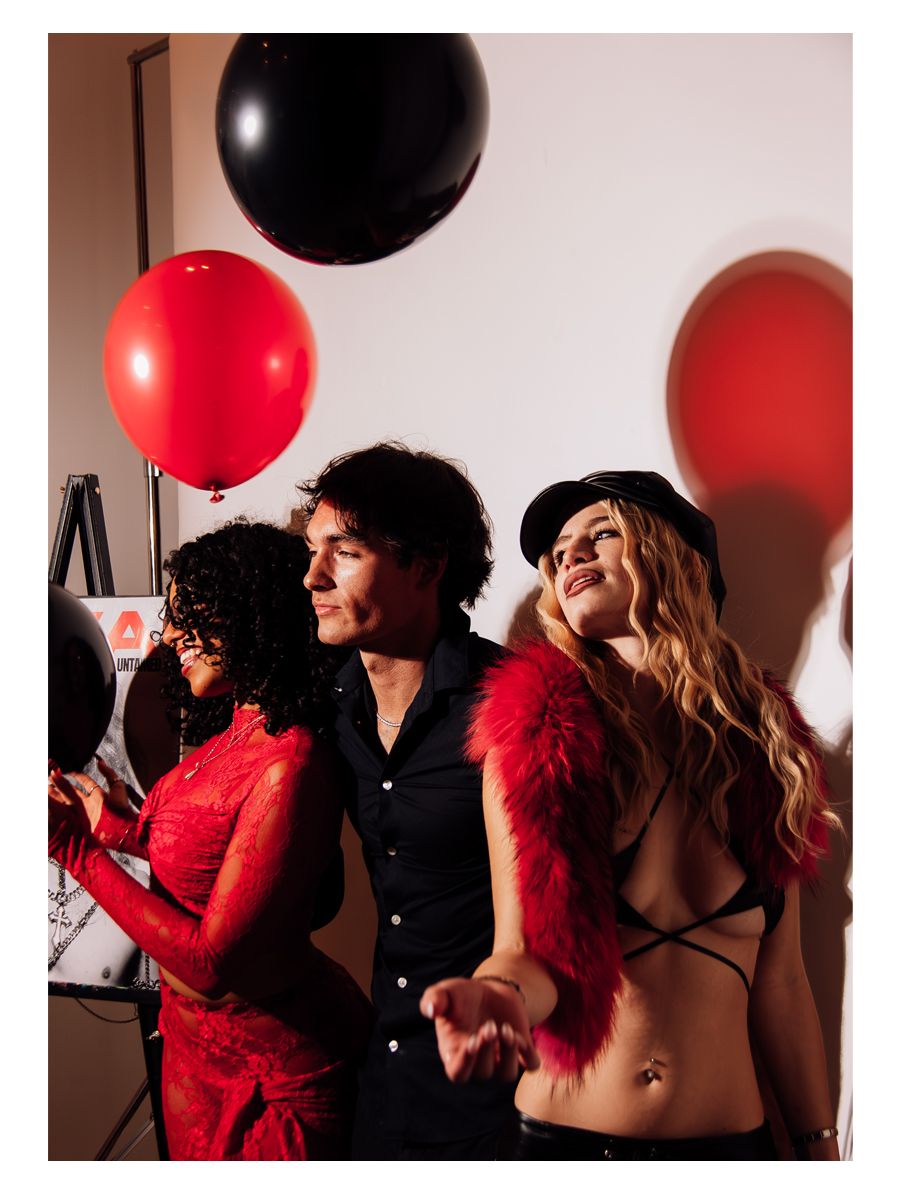 Three people laughing together at a party with red and black balloons, woman in red fur and leather cap stands out.