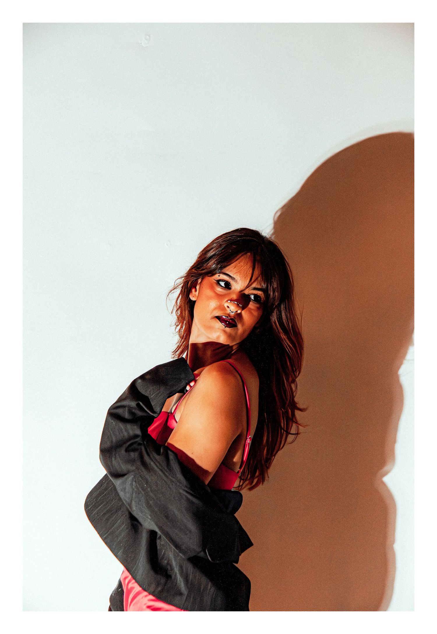 Woman in red satin dress and black jacket posing dramatically with shadow on wall.