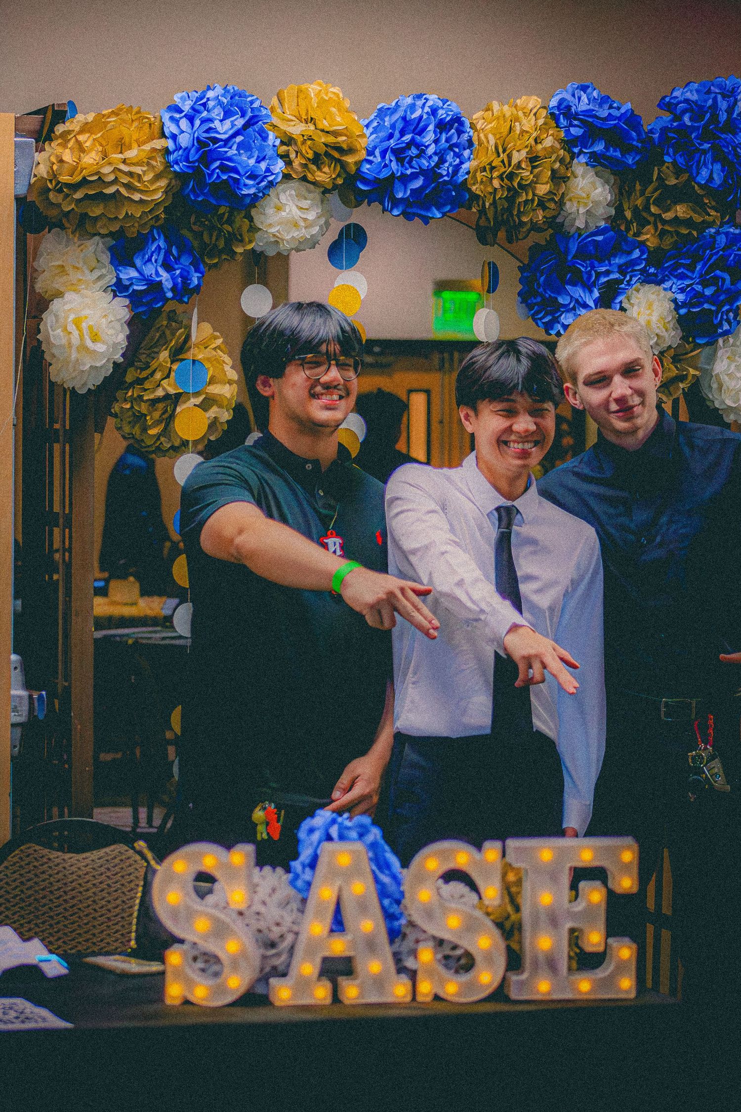 Three young men pose at a SASE event with blue, gold, and white paper flower decorations behind them.