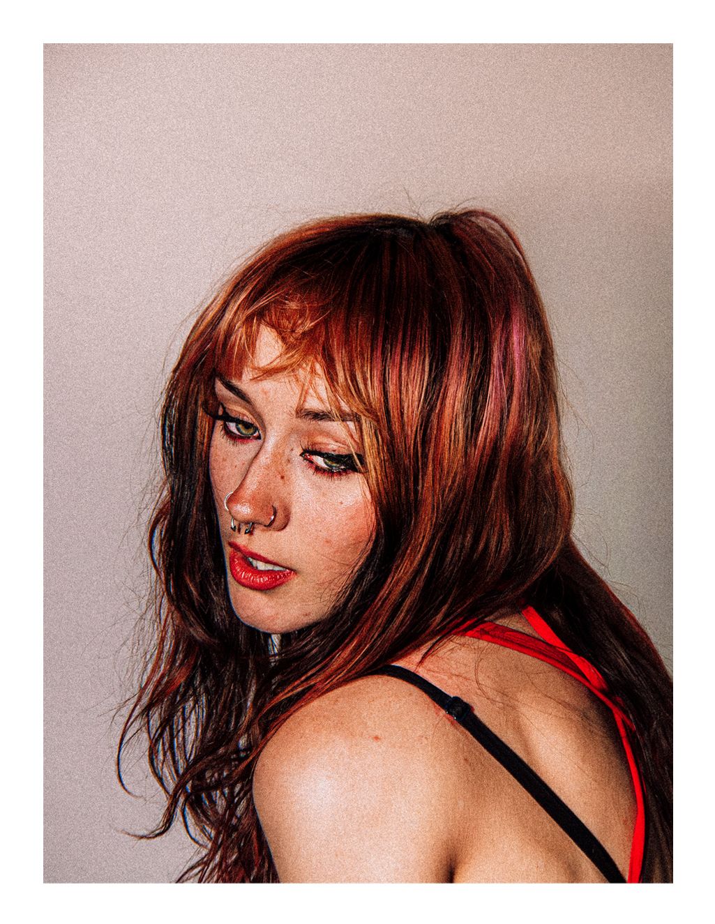 Side profile portrait of a young woman with auburn layered hair, red lips, and nose ring on neutral background.