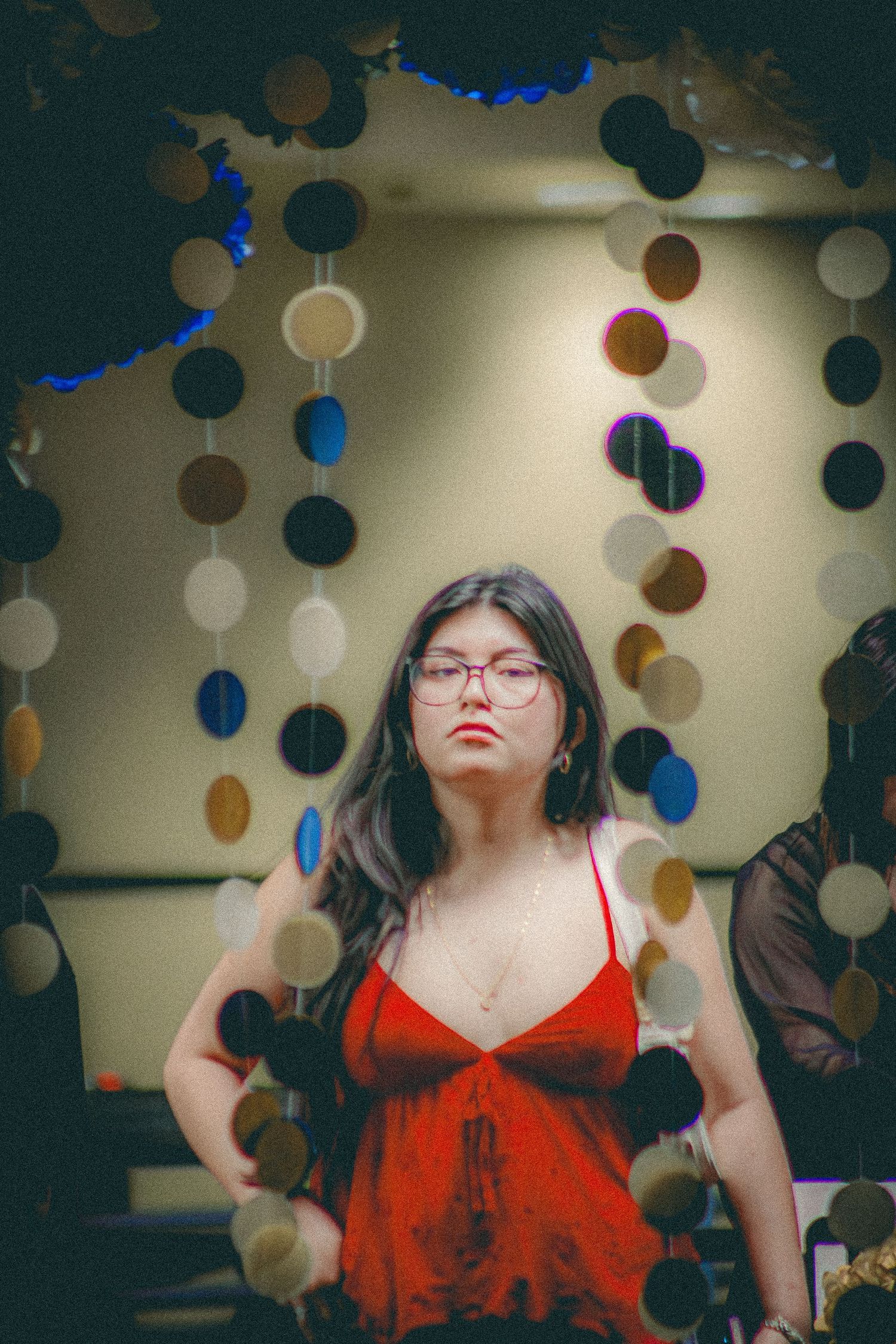 A young woman in a red dress with glasses poses in front of hanging circle garland decorations.