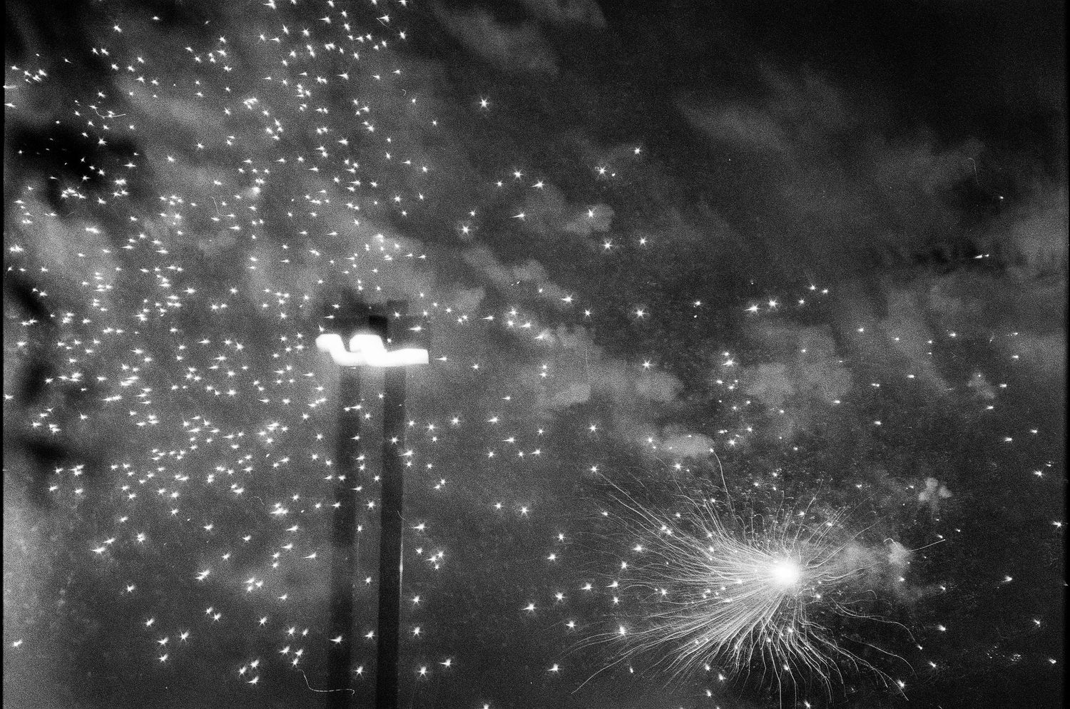 Black and white long exposure photograph of fireworks bursting in night sky near street lamp.