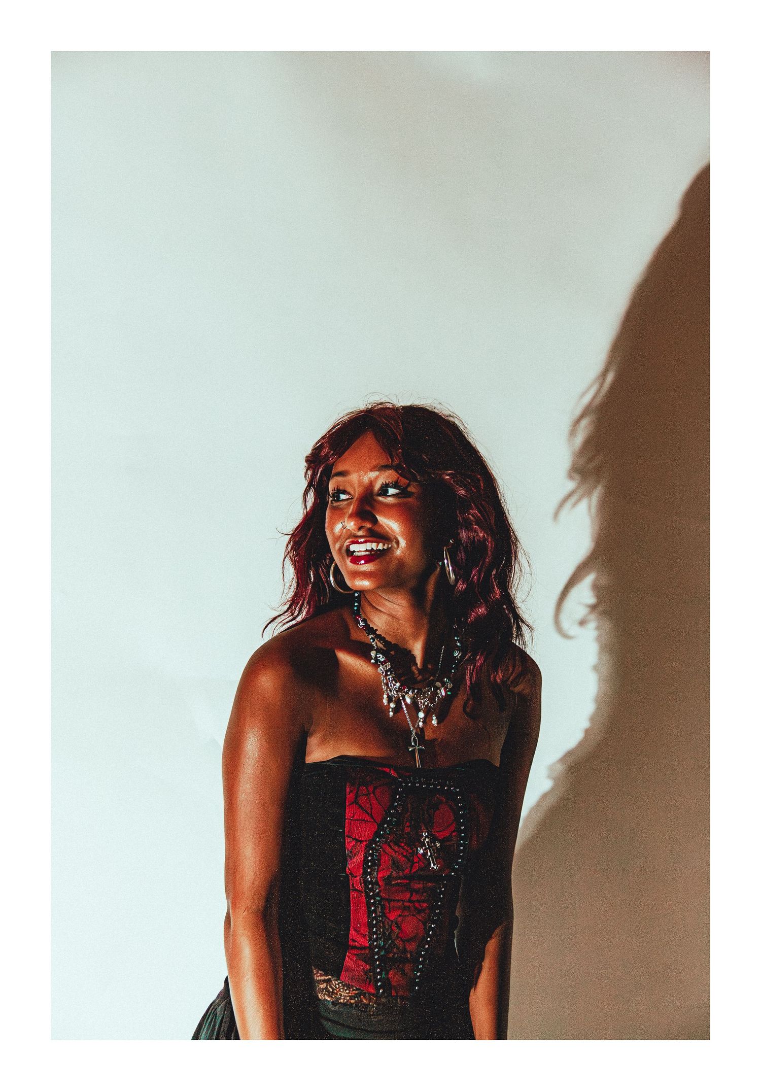Smiling woman in red corset top and layered necklaces poses in warm studio light.