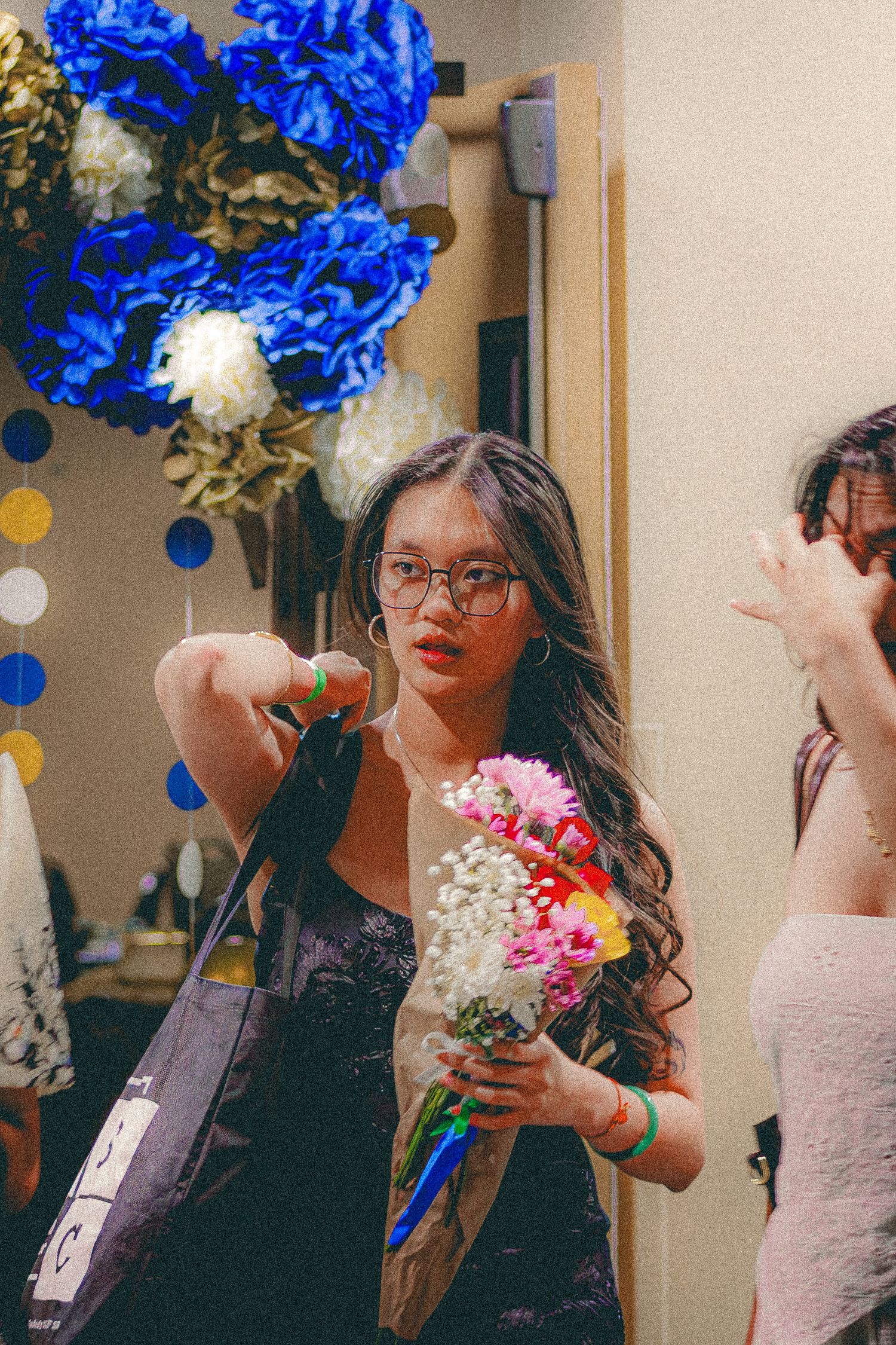 Young Asian woman with glasses holding colorful bouquet of flowers near blue paper flower decorations.