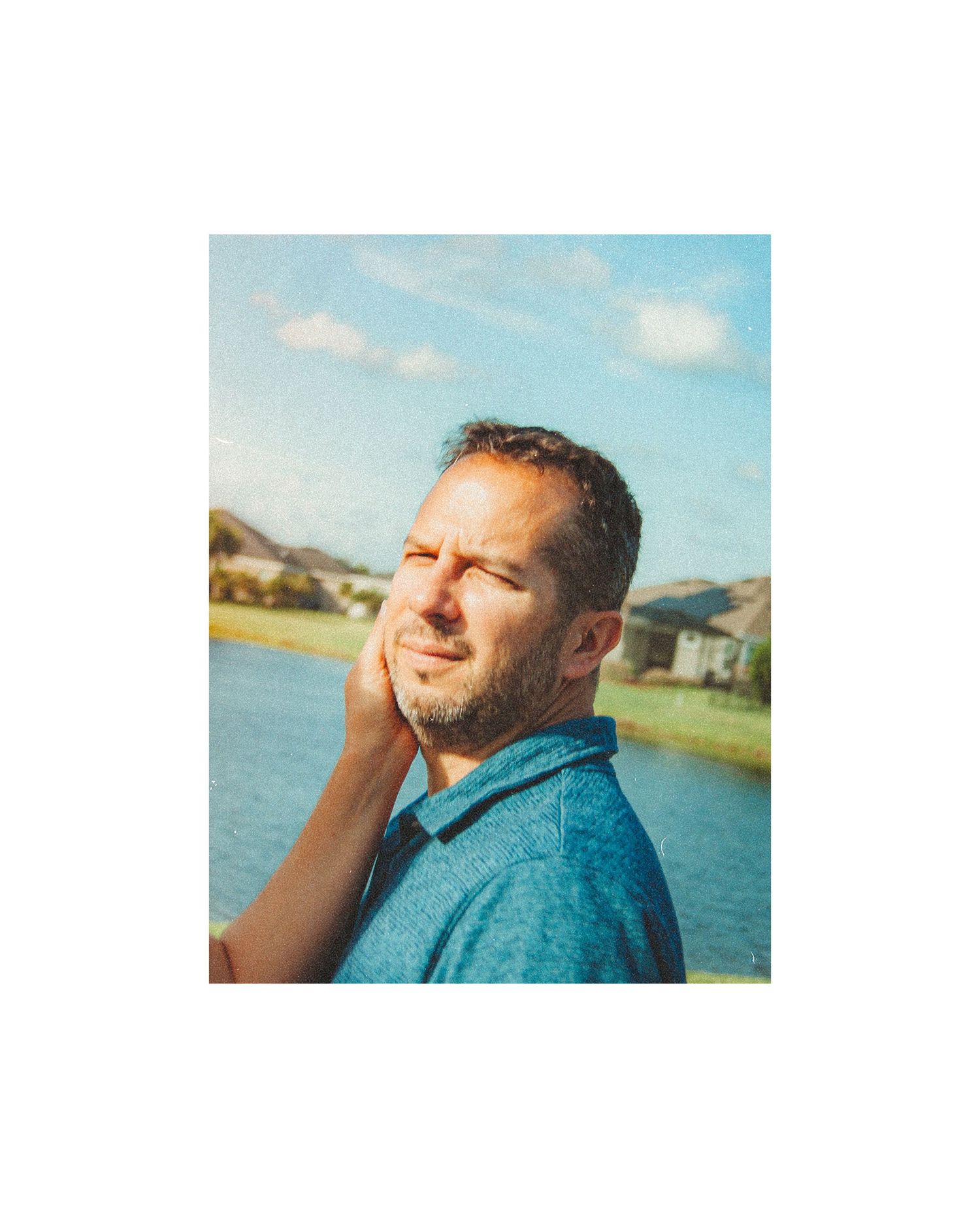A man in a blue polo shirt stands by a lake, hand on cheek, squinting in golden sunlight.