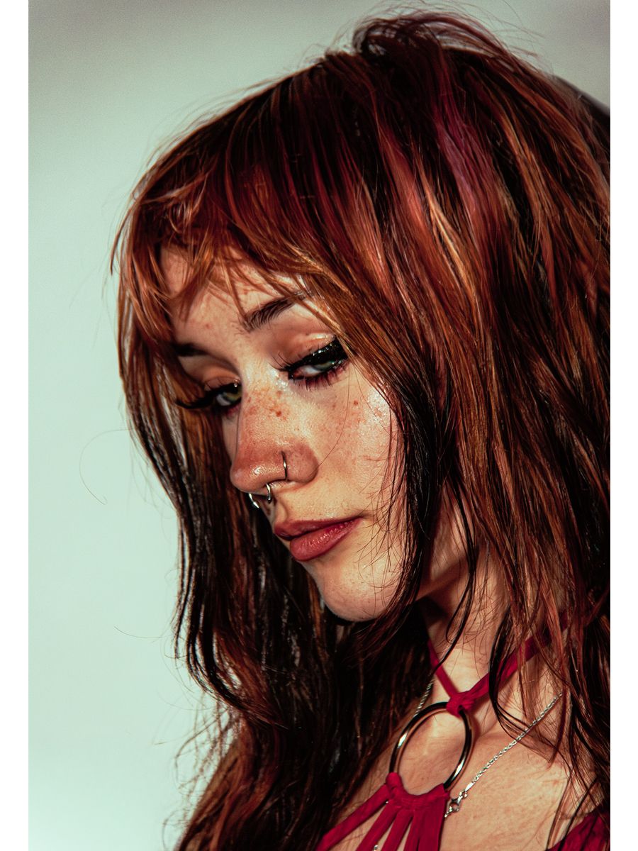 Close-up portrait of a young woman with auburn hair, freckles, nose ring, and dramatic eye makeup looking downward.