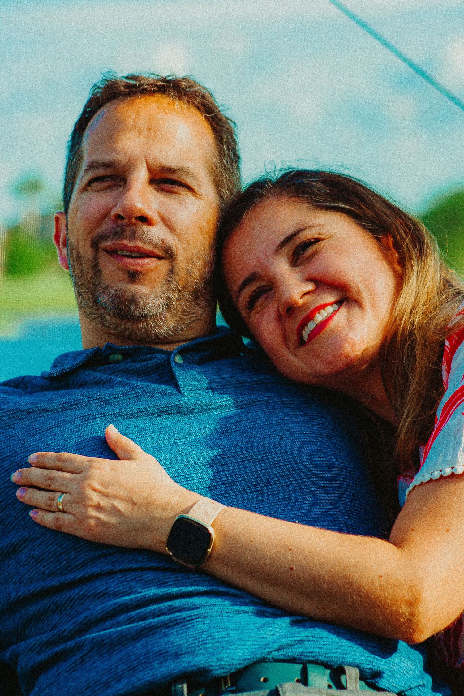 Happy couple embracing outdoors, woman smiling and hugging man in blue shirt, both wearing rings, sunny day.