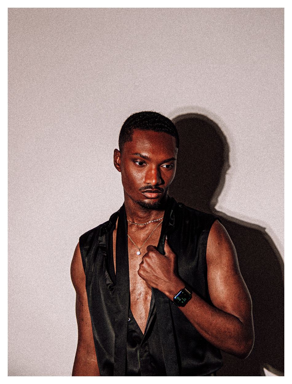 A young Black man in a black sleeveless shirt poses confidently wearing gold chain necklaces.