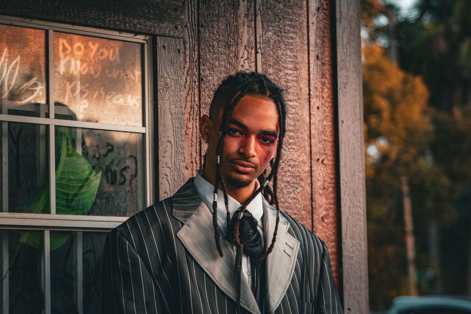 A young man with dreadlocks in a pinstripe suit leans against a weathered wooden wall outdoors.