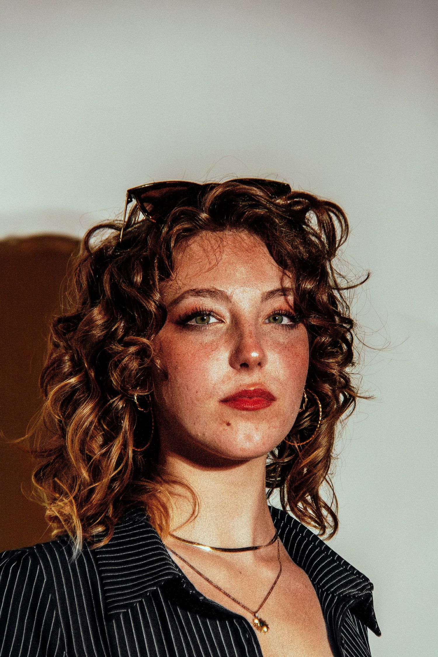 Young woman with curly auburn hair, freckles, red lips, hoop earrings, and a striped shirt poses confidently.