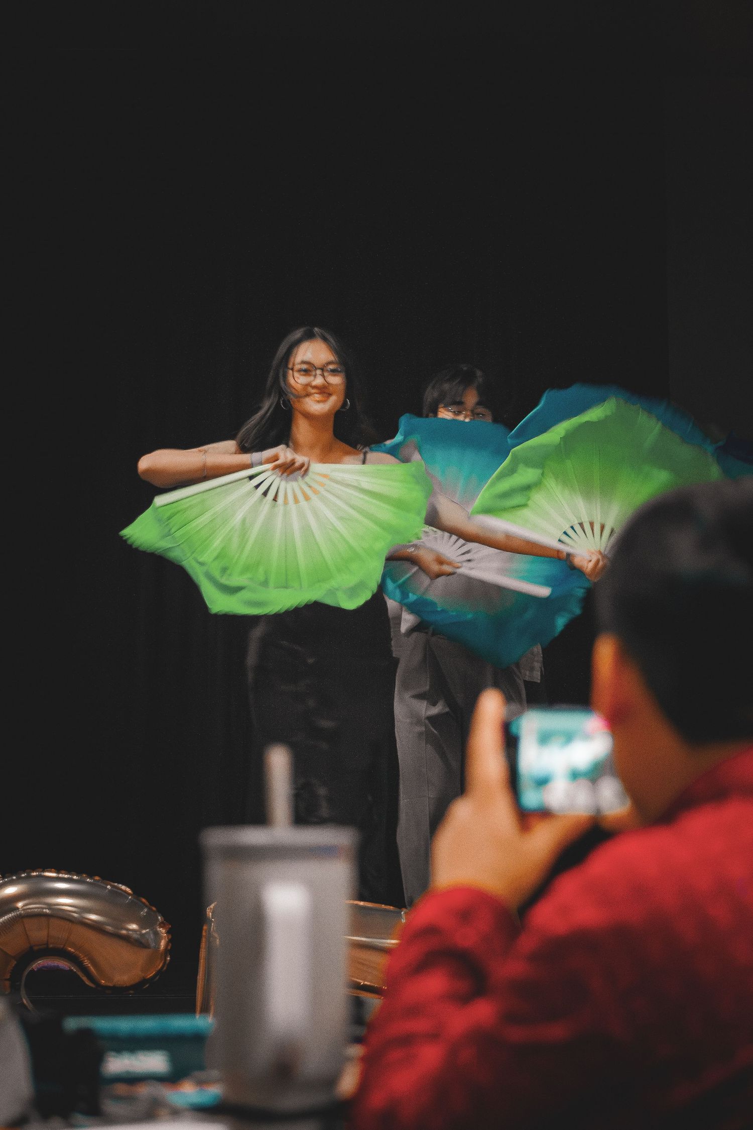 Smiling young Asian woman performing fan dance with green silk fans while audience member photographs her.