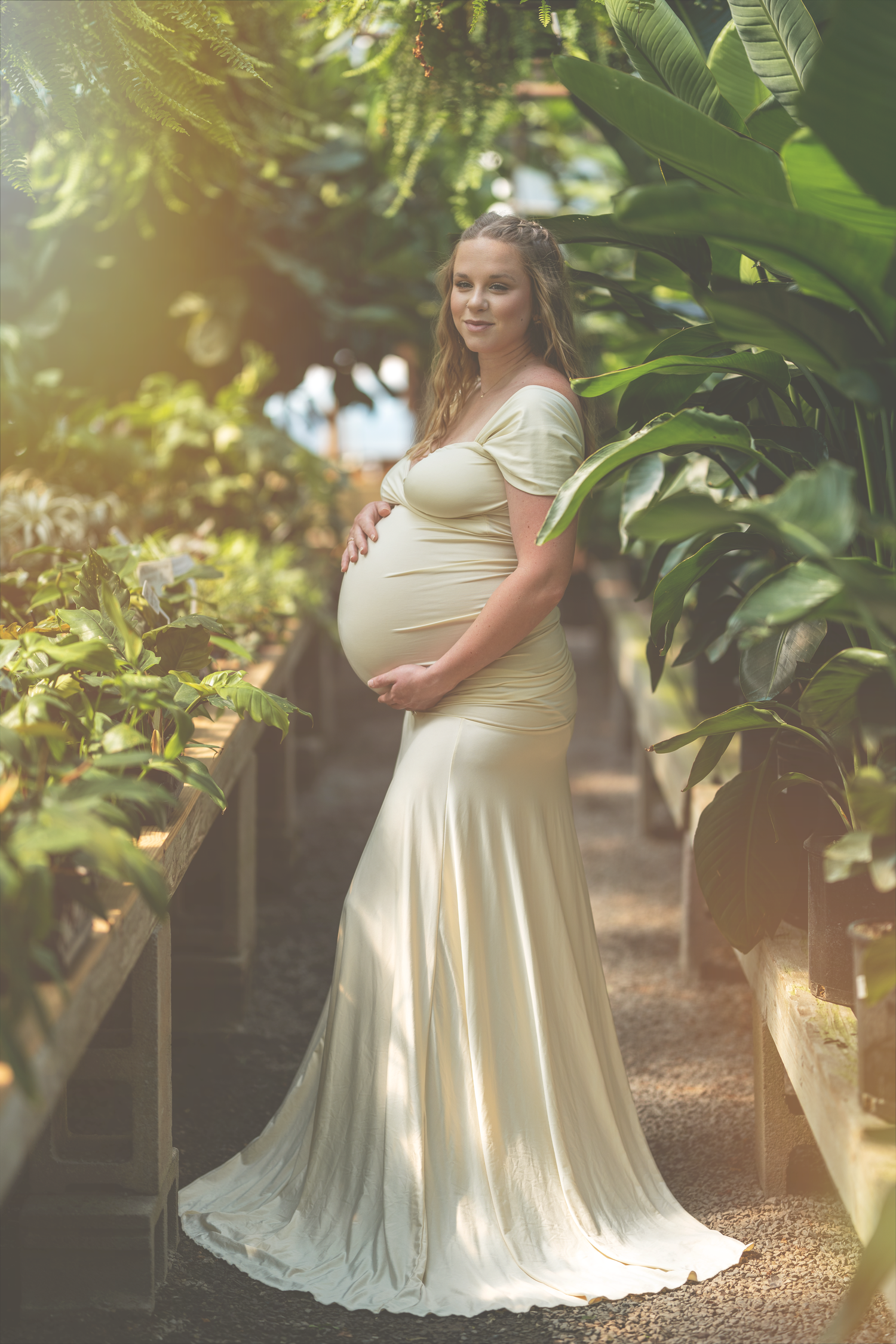 A pregnant person in a flowing cream maternity dress poses in a backyard garden setting with dappled sunlight filtering through.