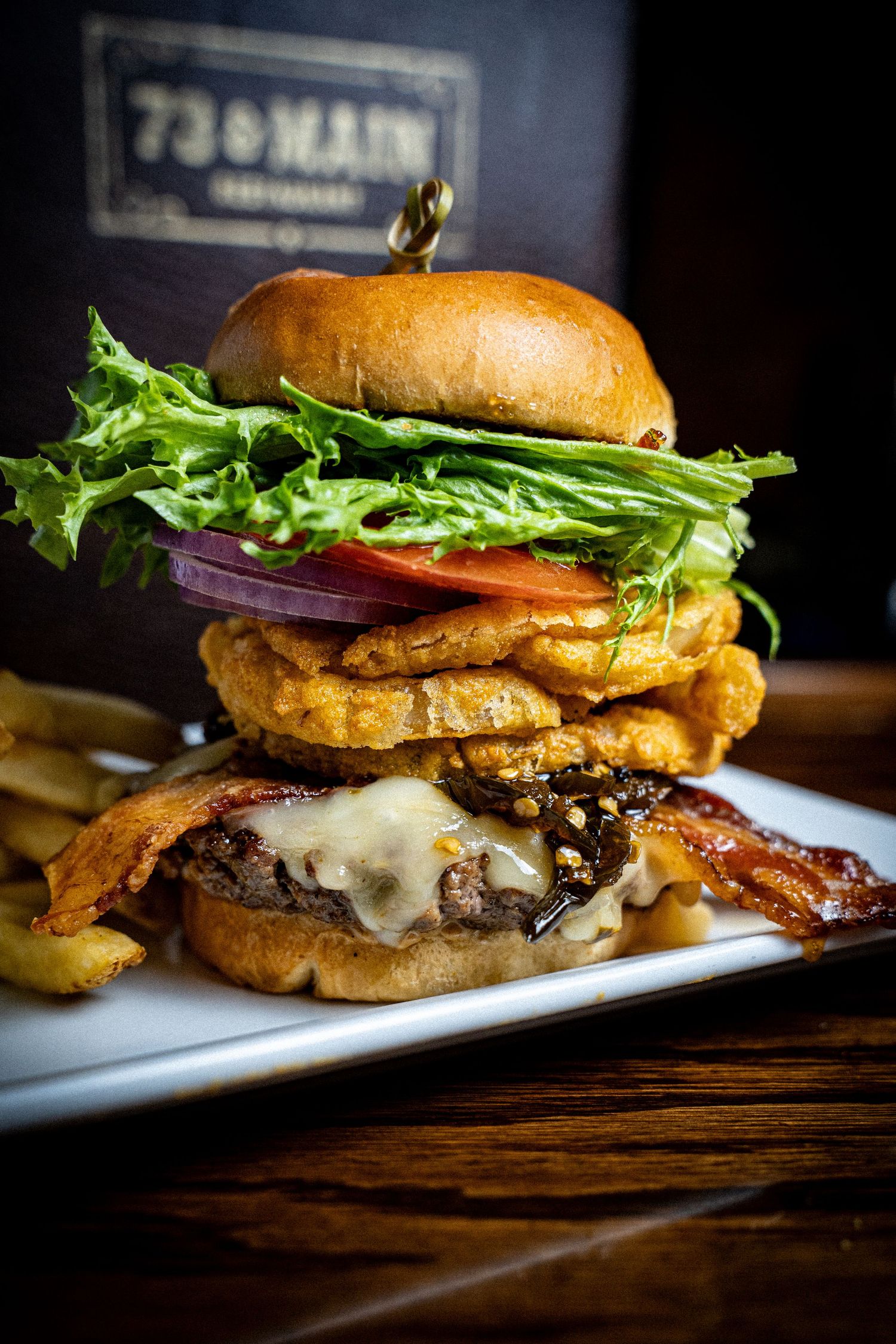 Gourmet burger stacked high with bacon, lettuce, and tomato served with fries on a white plate.