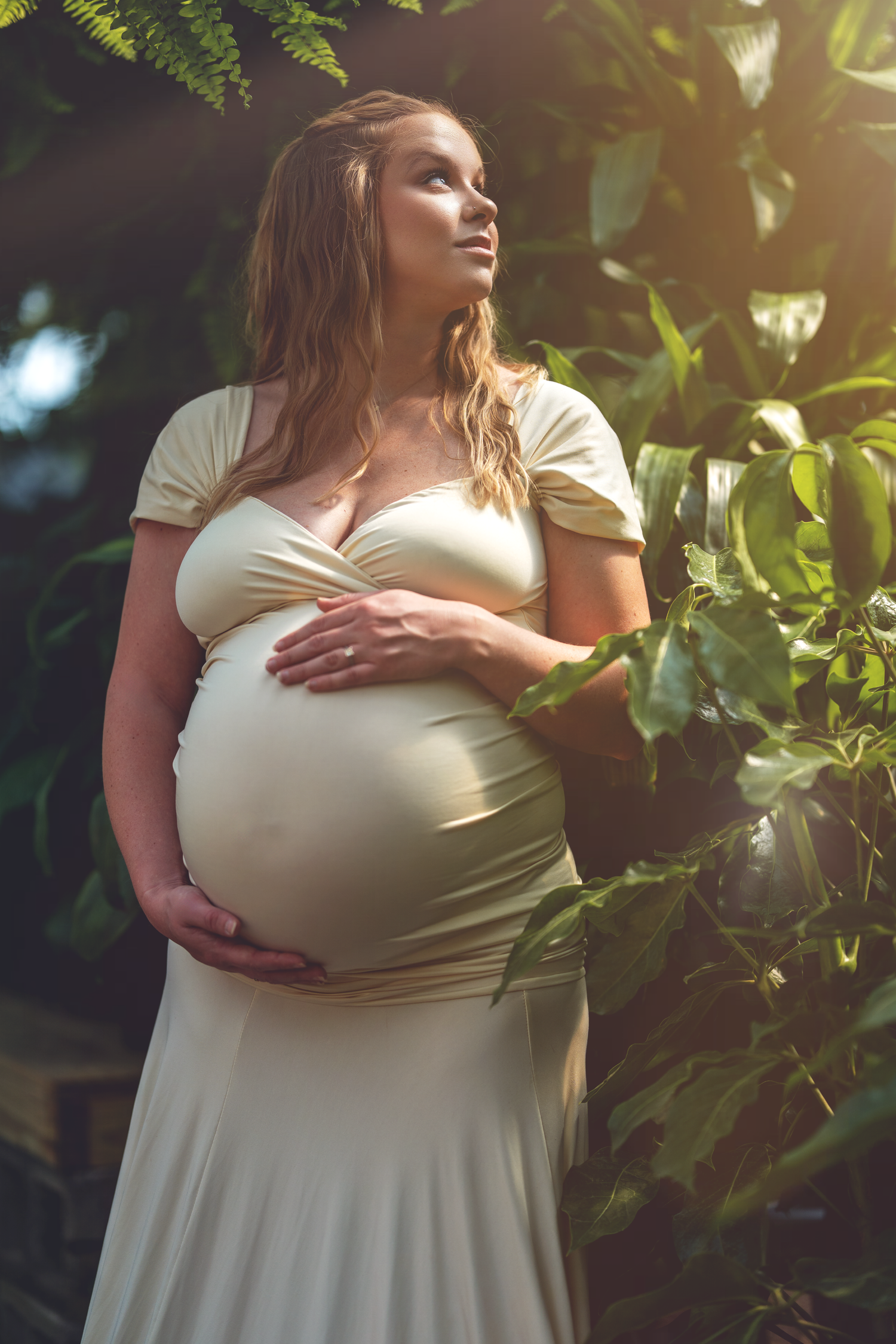 Expectant woman in white dress stands in dappled sunlight among lush garden foliage while cradling her baby bump.