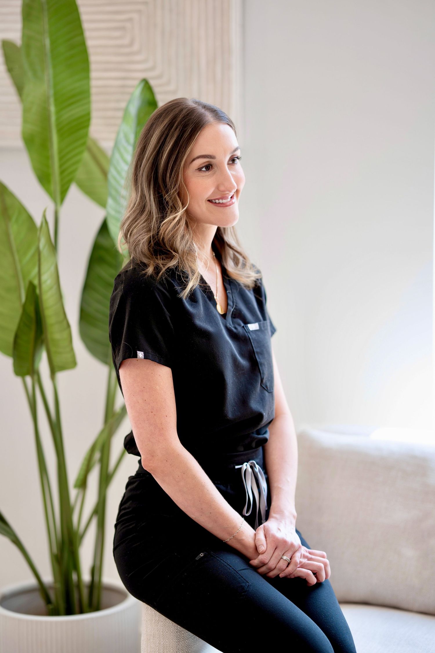 Healthcare professional in navy blue scrubs sits casually near a large tropical plant in a bright modern office setting.