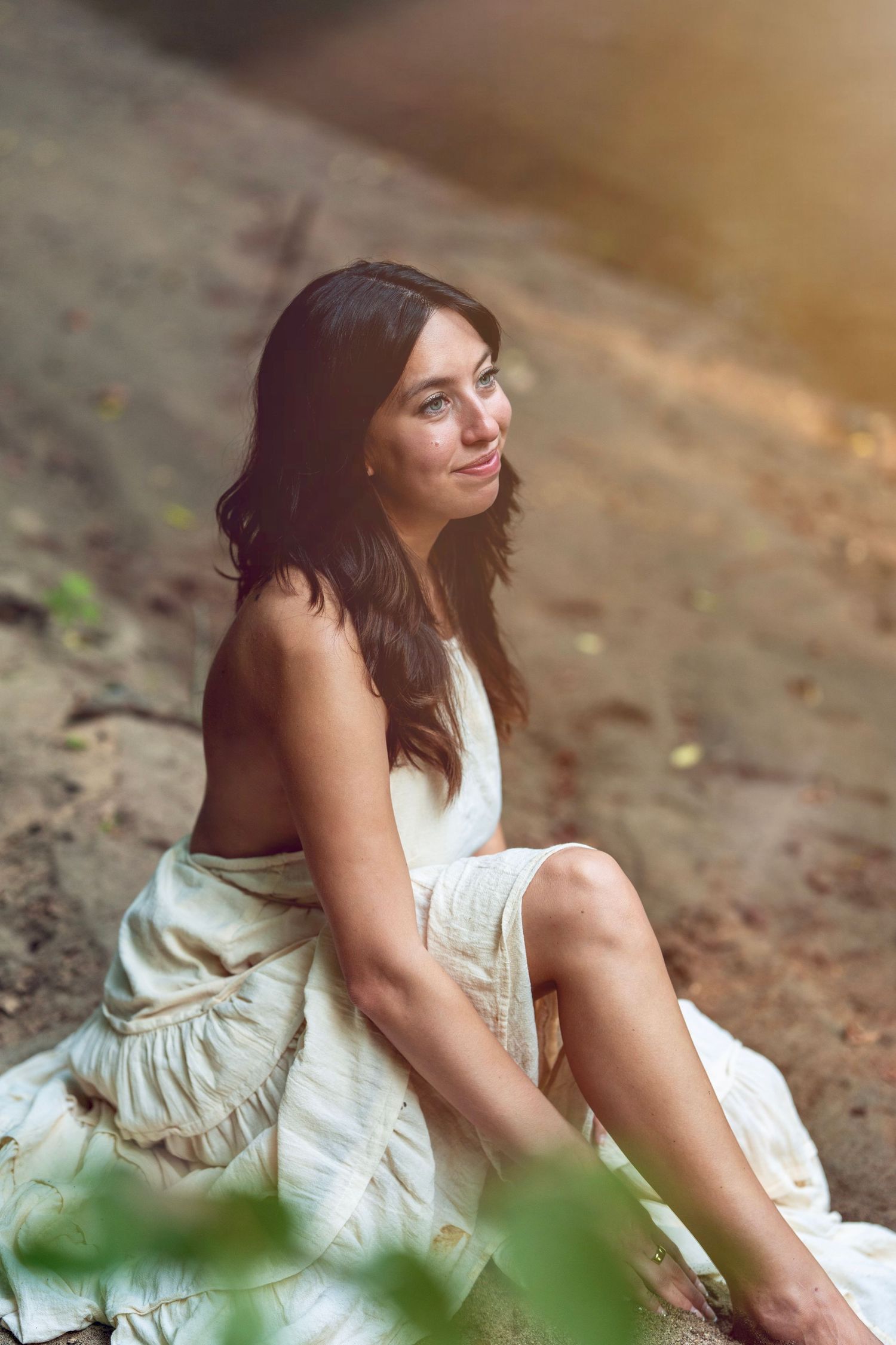 Young woman in white dress sits on ground outdoors during sunset with dreamy warm lighting and blurred foliage.