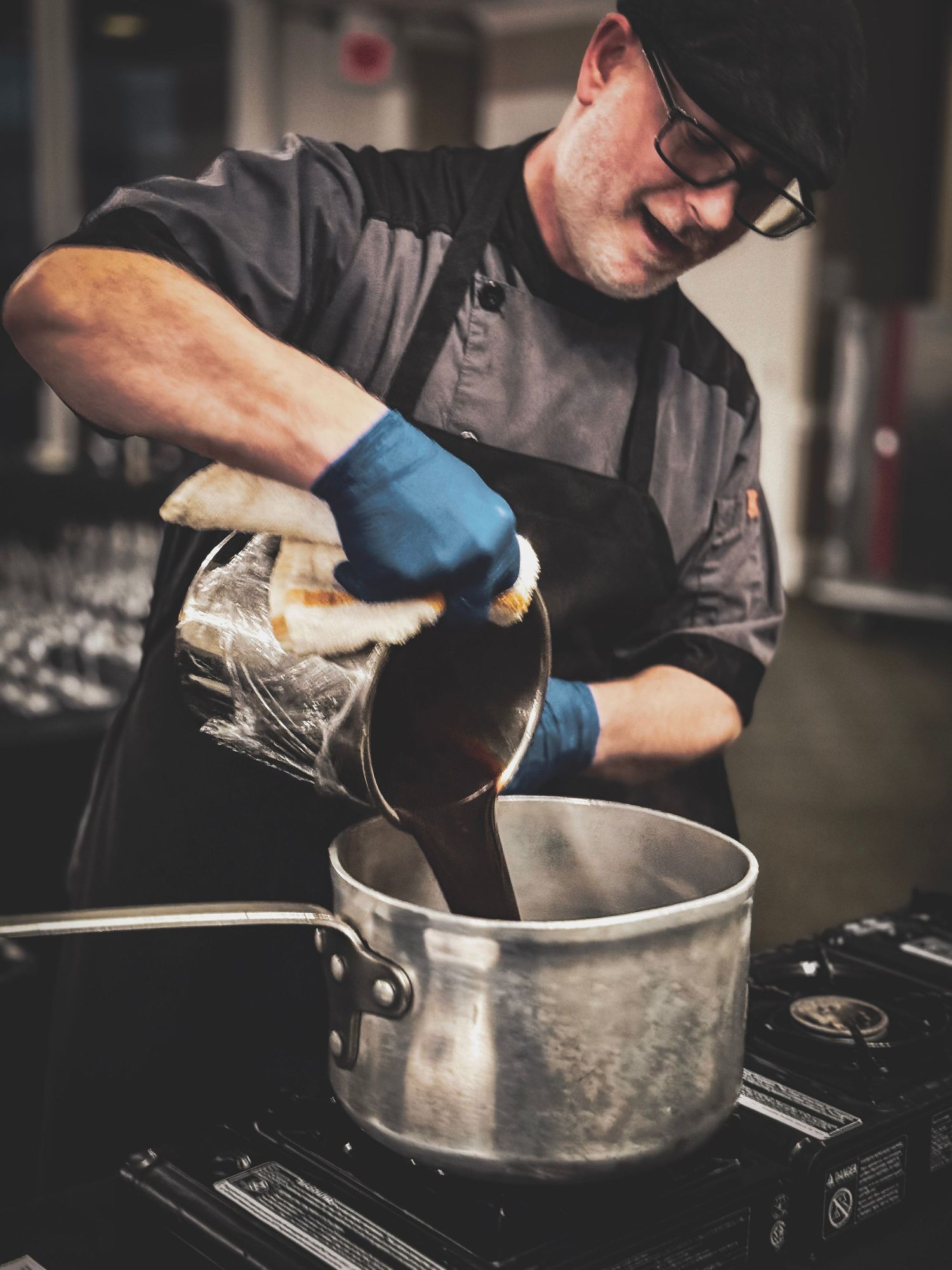 A chef wearing blue gloves pours liquid from a metal container into a pot in a professional kitchen.