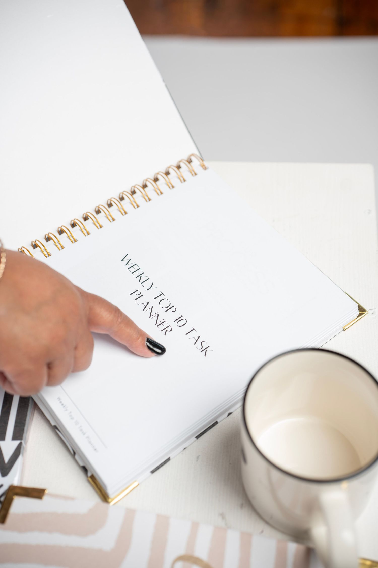 A white spiral notebook and silver coffee mug on a desk with someone's hand holding a pen to write.
