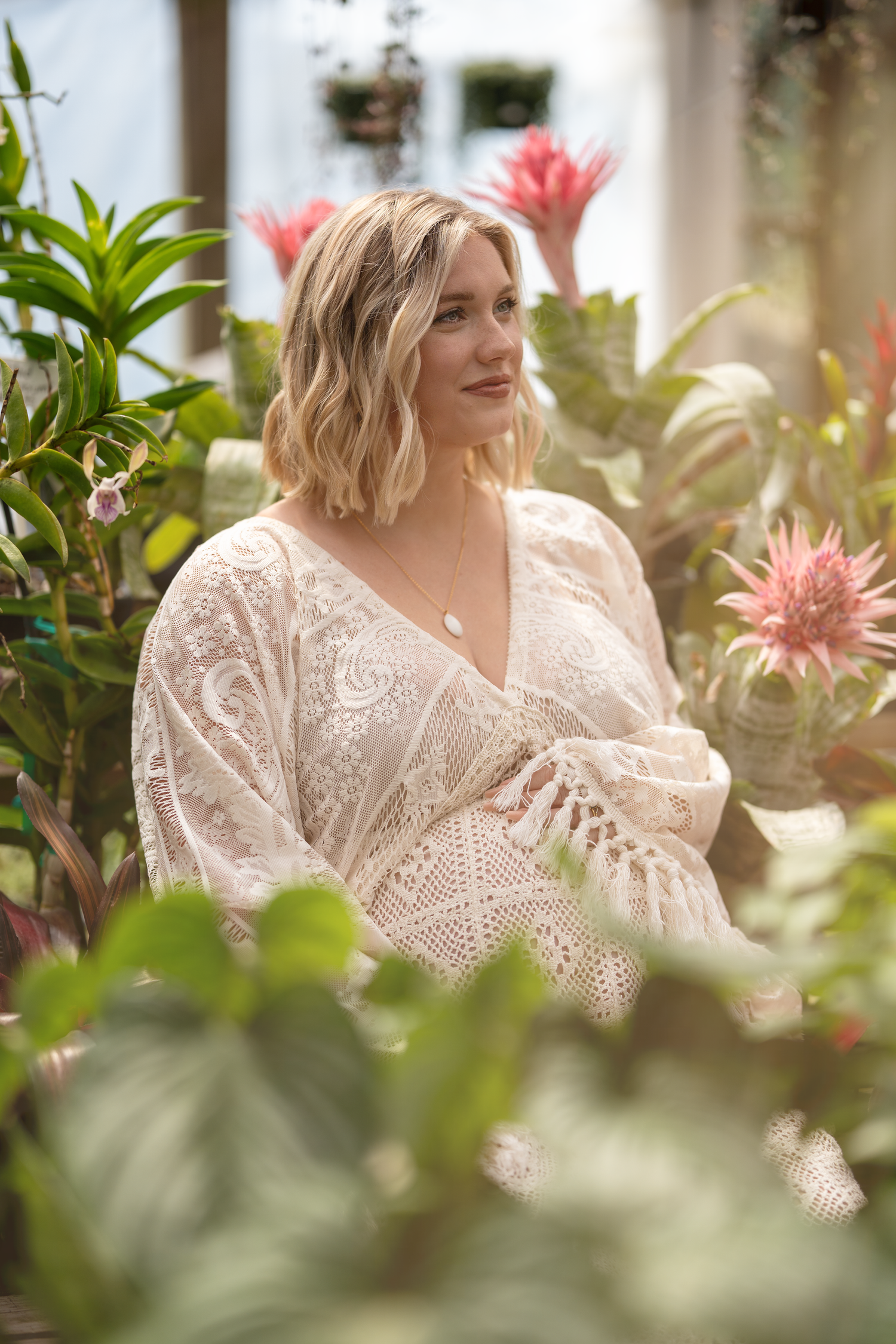 A person wearing a romantic white lace dress poses in a garden setting surrounded by pink flowers and greenery.