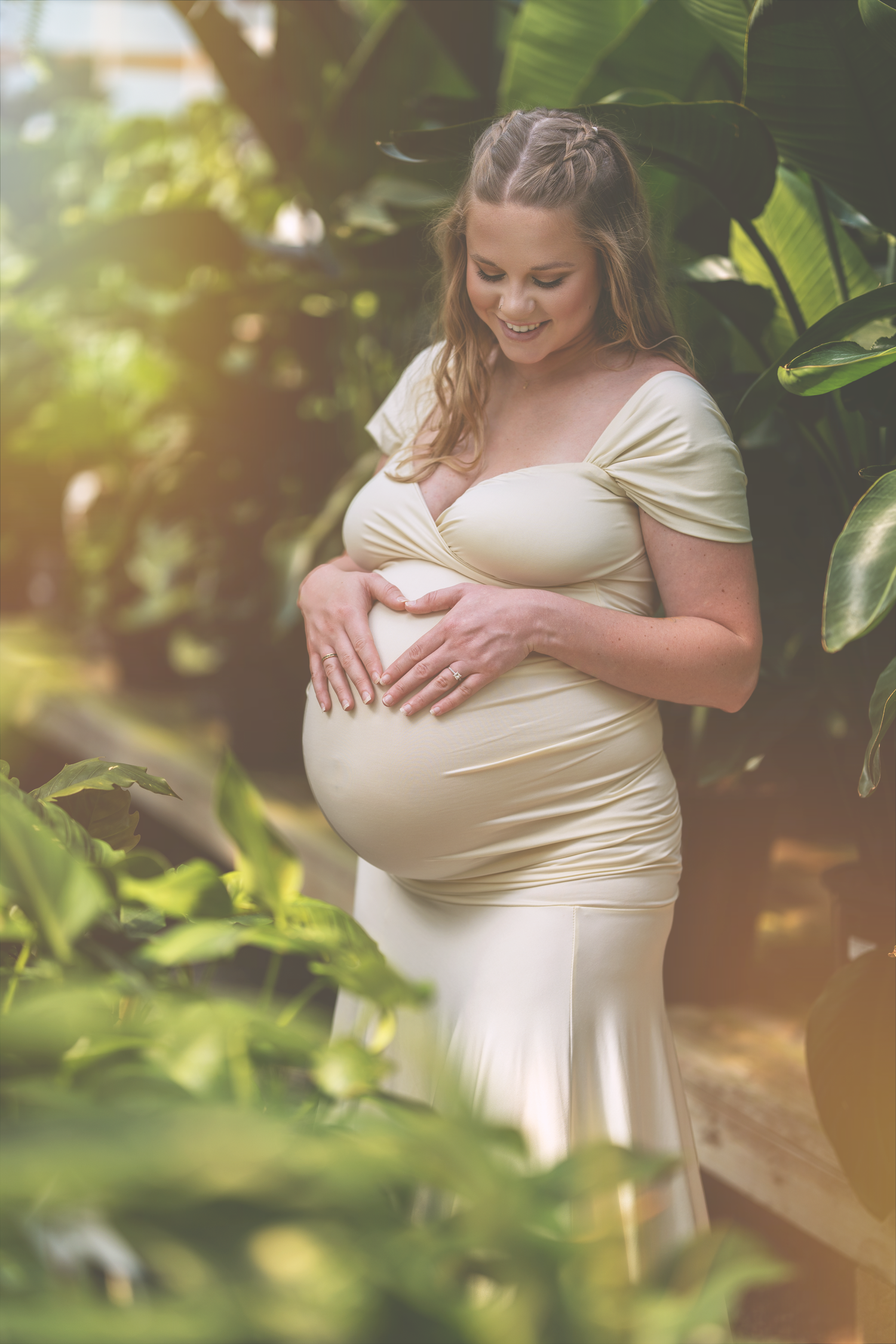 Expectant mother in a cream-colored dress poses in a garden setting with natural sunlight filtering through foliage.