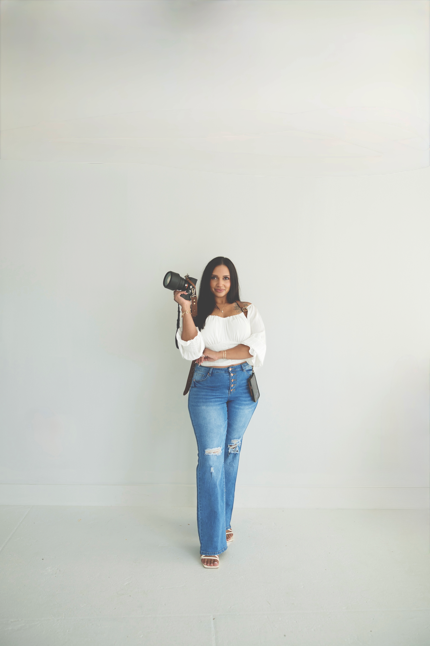 A photographer in ripped jeans and white crop top poses with a professional camera against a gray backdrop.