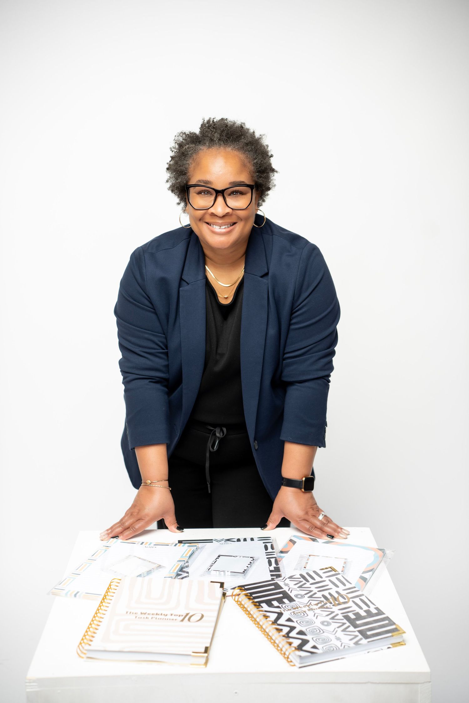 Professional in a navy blazer and glasses leans over a desk with various documents spread out on white surface.