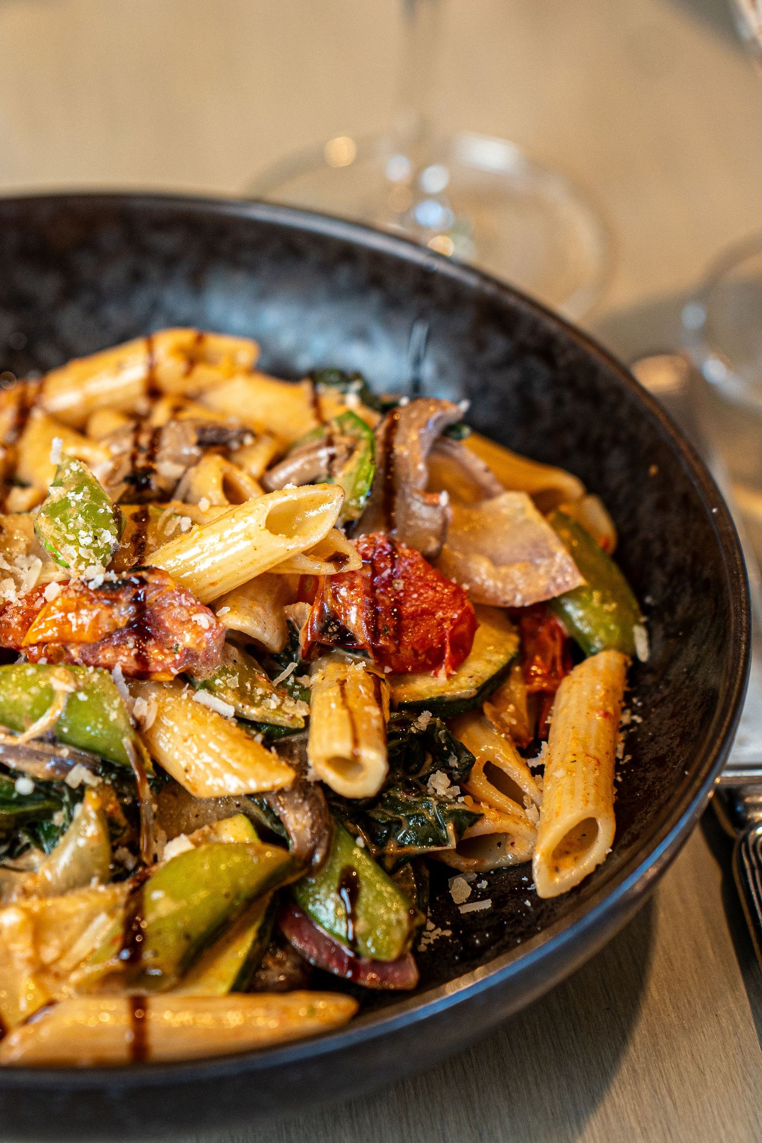 A rustic bowl of penne pasta with roasted vegetables and mushrooms in a dark ceramic dish.