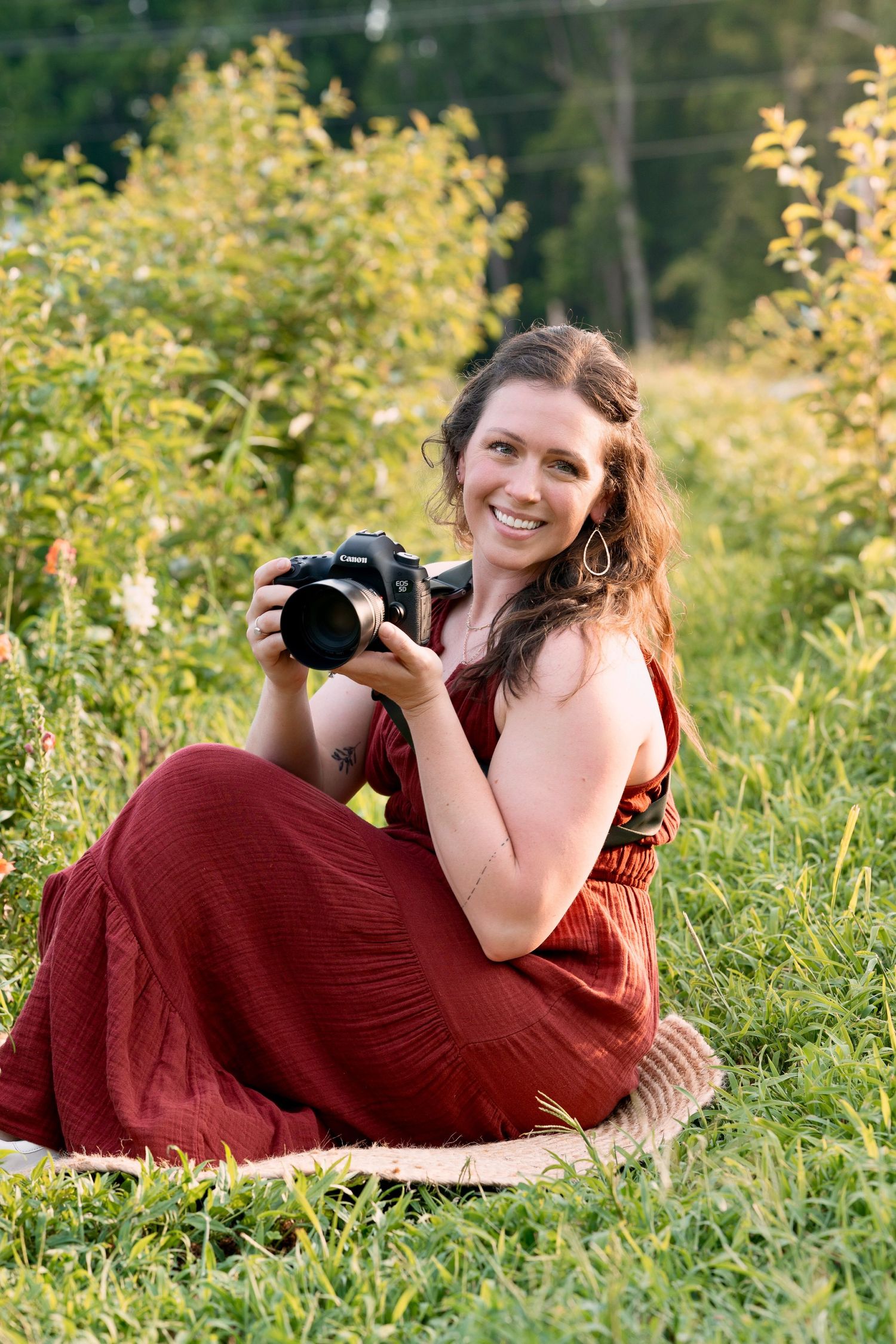 Photographer in red dress sitting in field with yellow flowers holding camera.