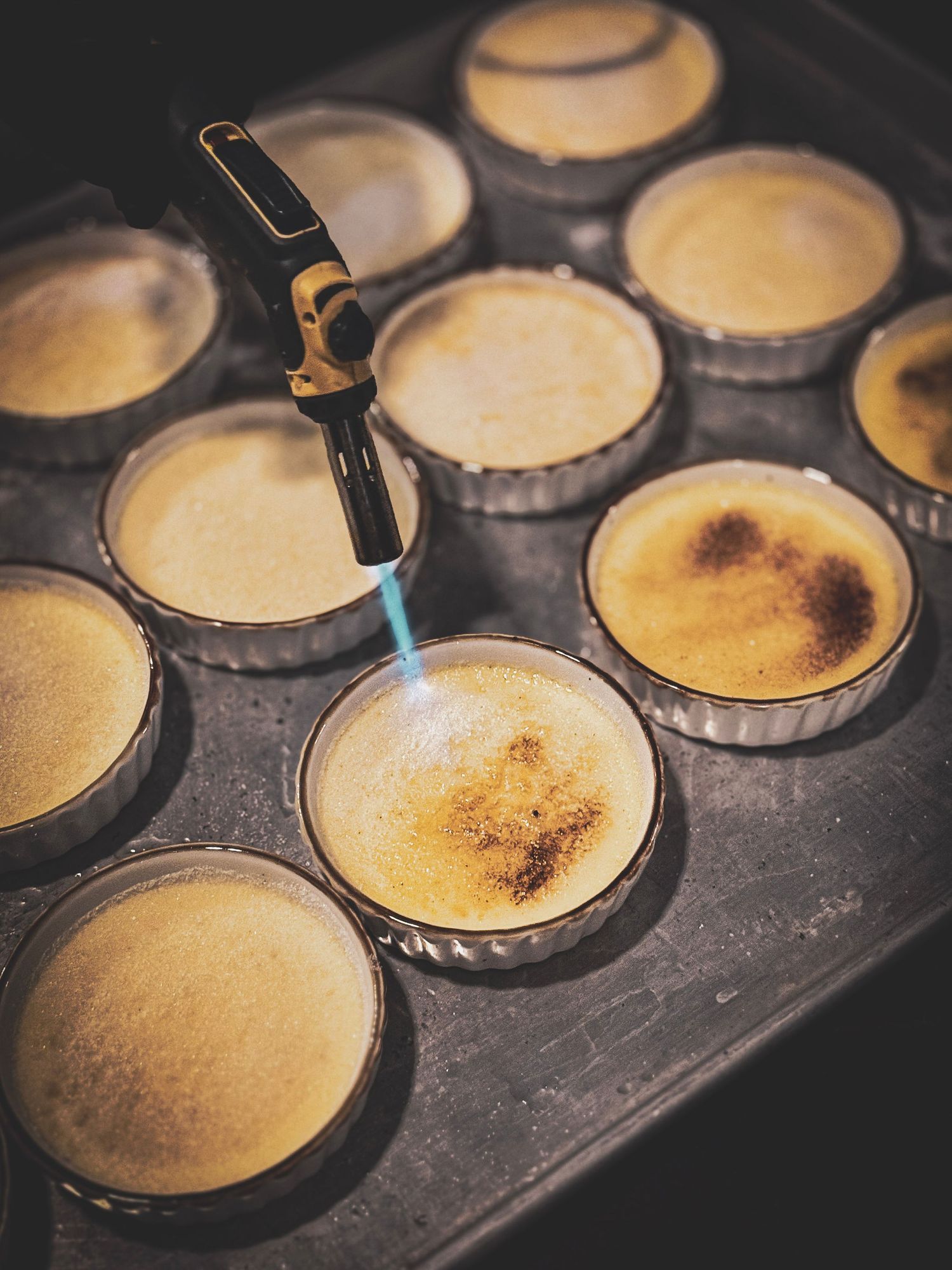 Culinary torch browning the tops of custard tarts in a metal baking pan.