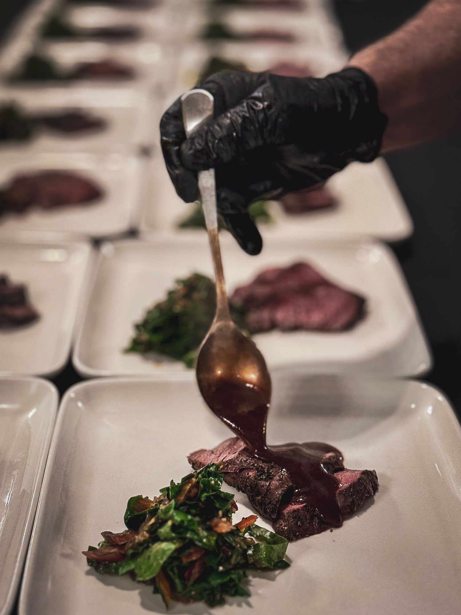 Chef in black gloves pouring sauce over sliced meat and greens on white plates.