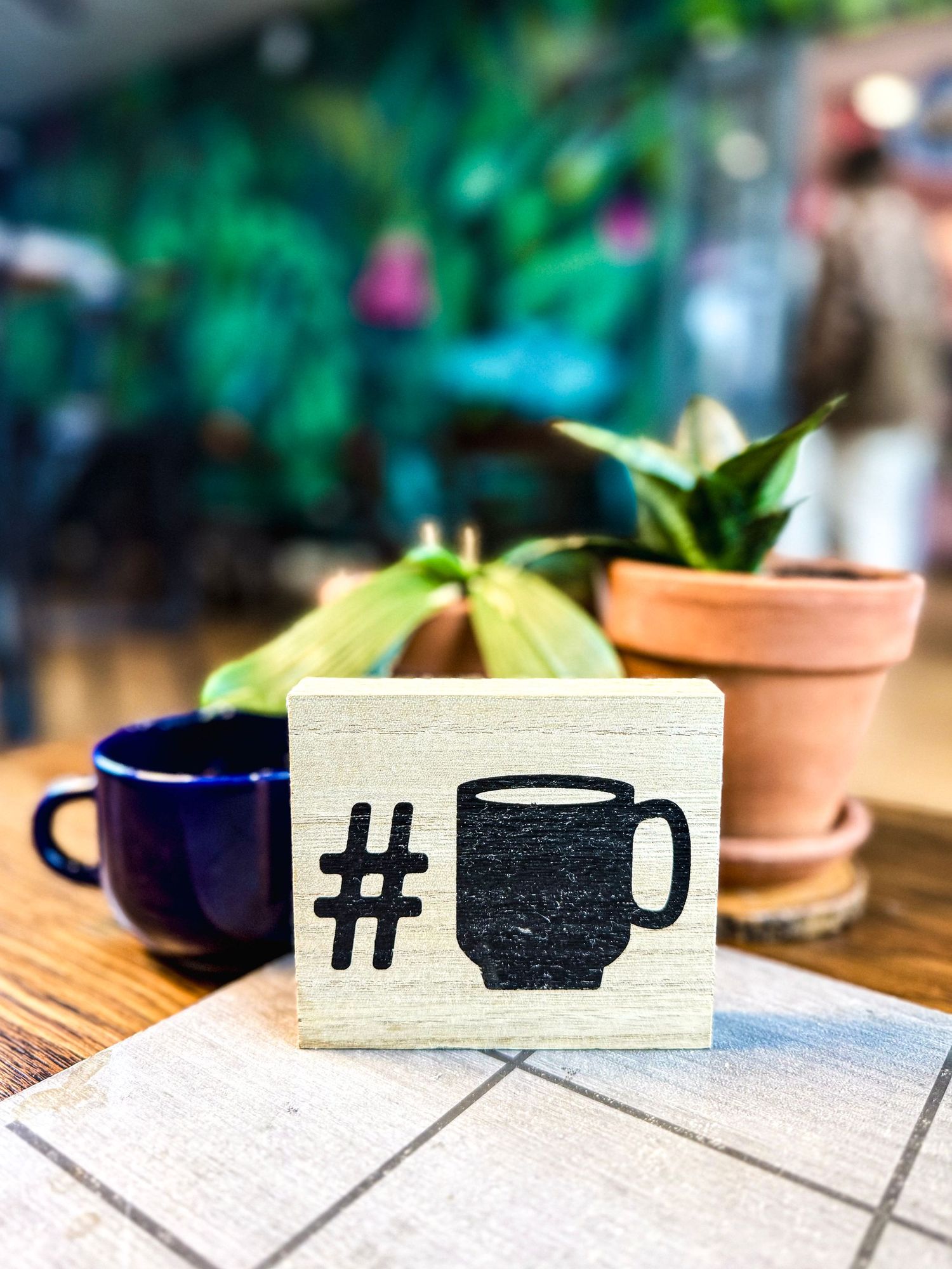 Decorative coffee hashtag sign with blue mug and small potted plants against blurred green background.