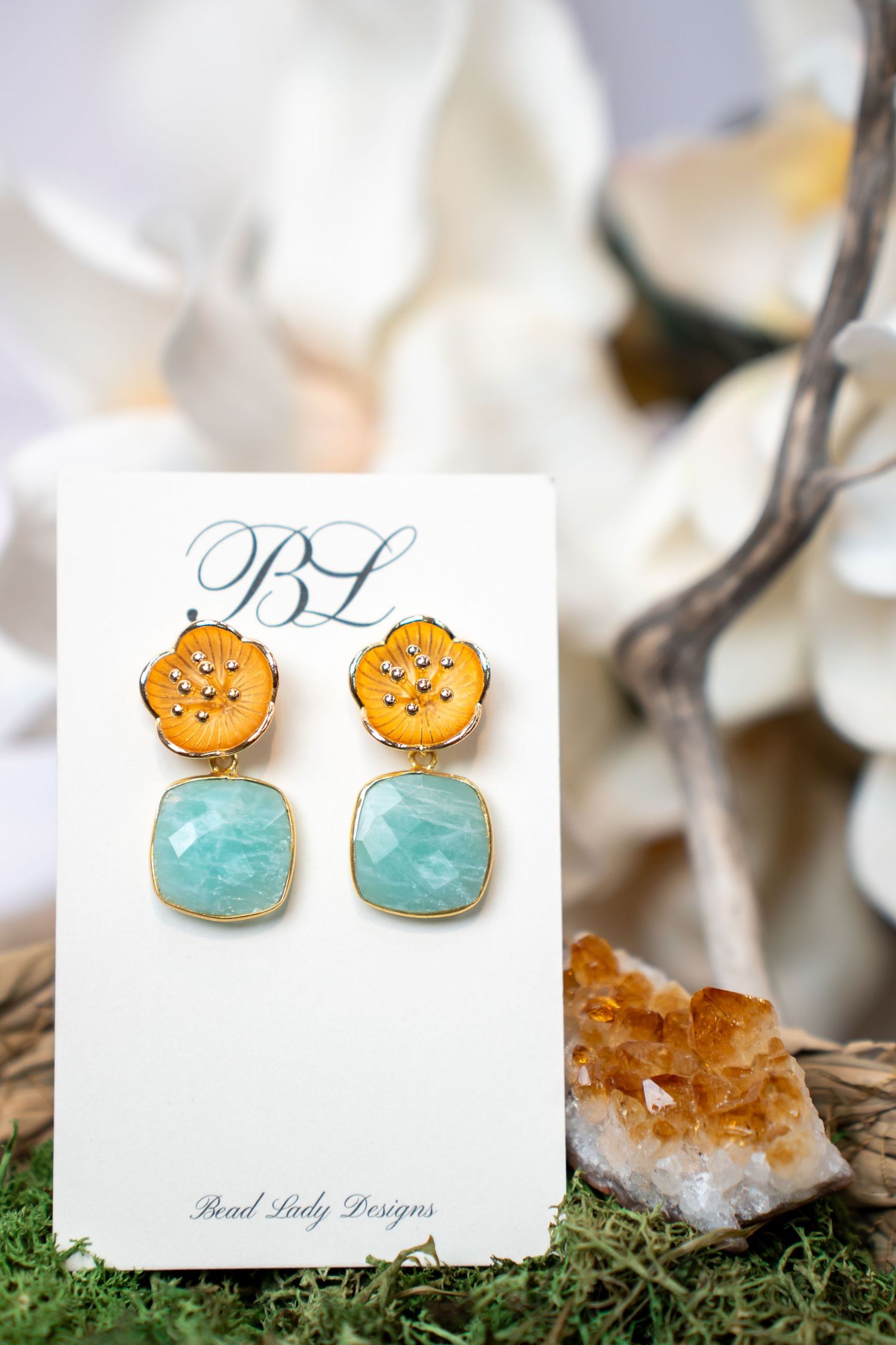 Gold flower earrings with turquoise stones displayed on a branded card with crystals and white flowers.