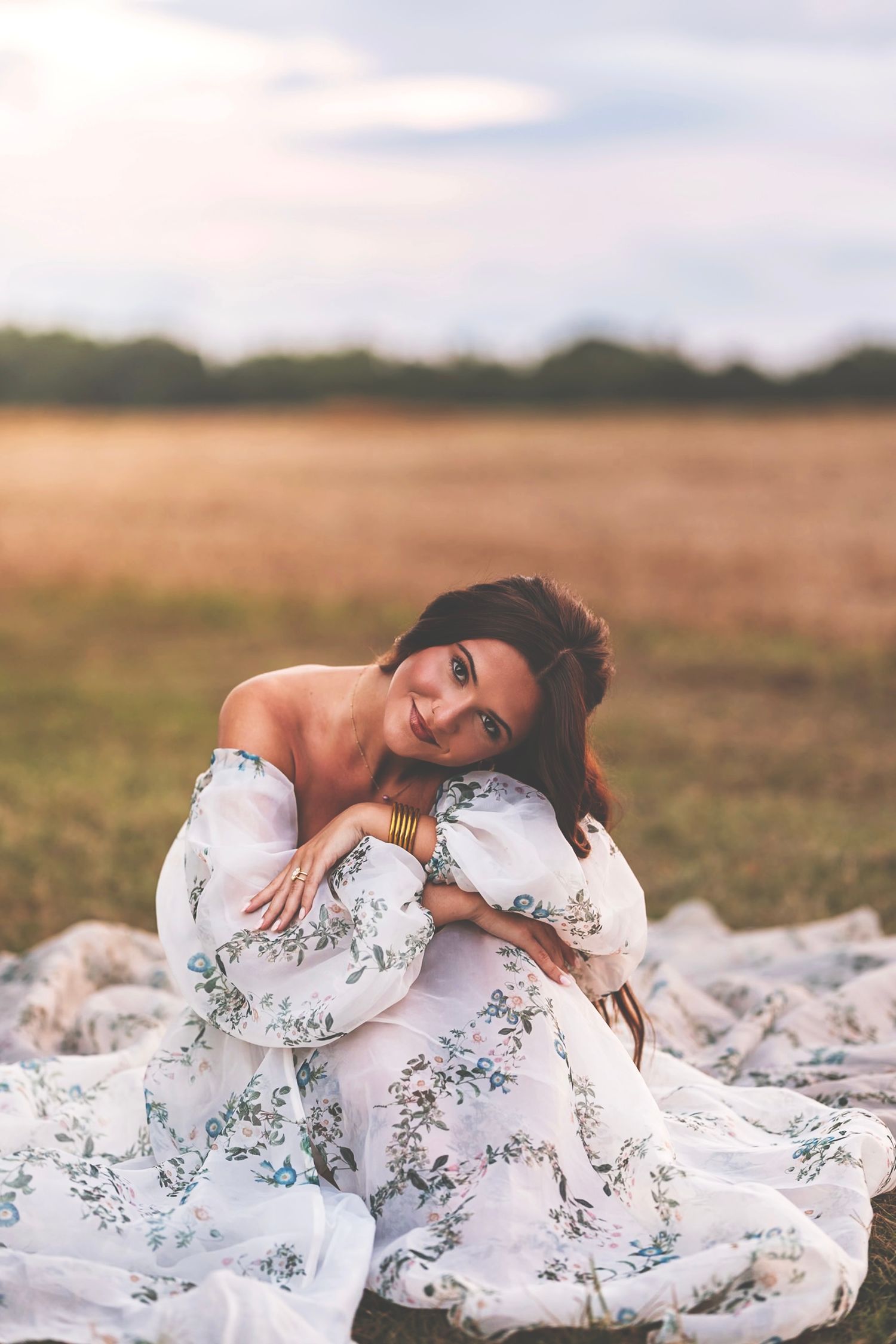 A woman in a flowing white floral dress lies gracefully on a grassy field during golden hour.