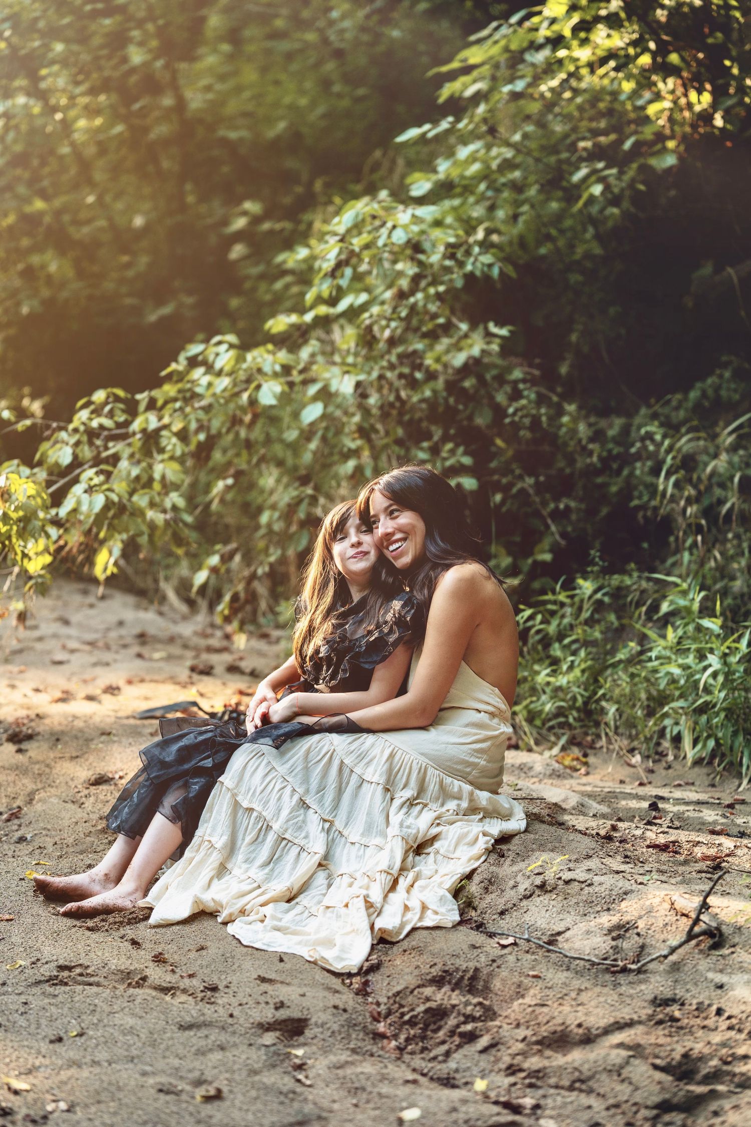 Two people in flowing white dresses sitting together on stone steps surrounded by greenery in warm sunlight.