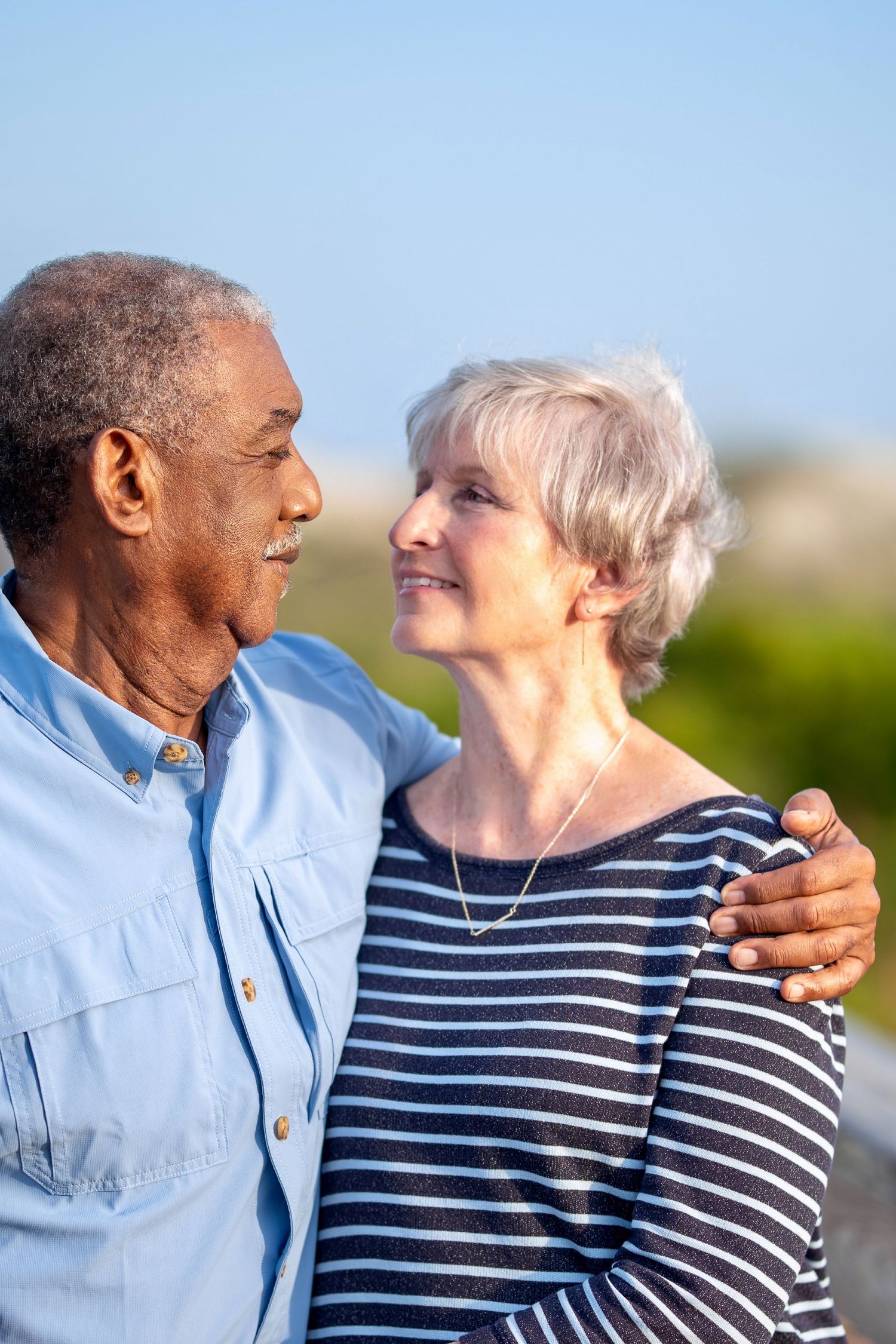An elderly couple embraces outdoors on a sunny day, with one wearing a blue shirt and the other in a striped top.