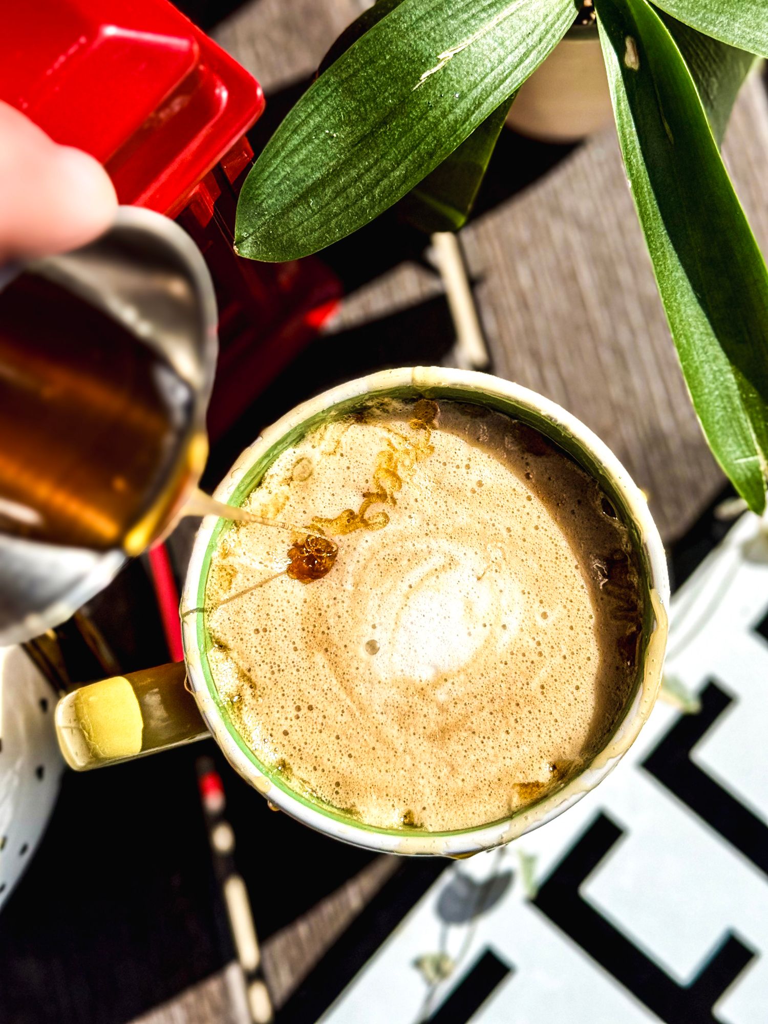 Overhead view of a frothy latte in a gold cup with green plant leaves visible.