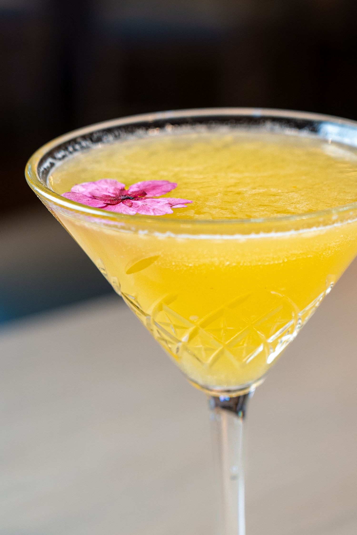 Yellow martini cocktail garnished with a pink flower in a sugar-rimmed glass.