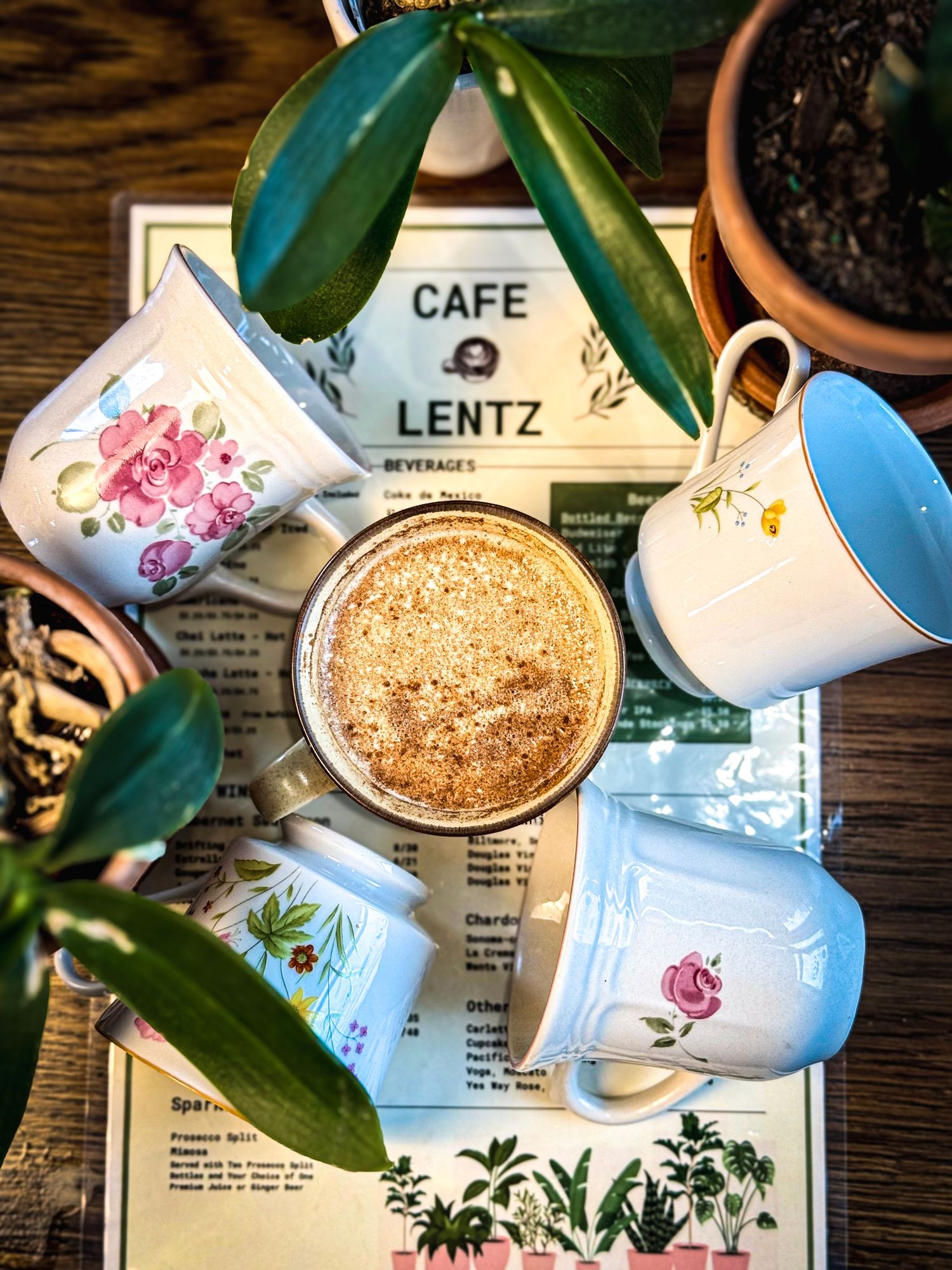 Vintage teacups and cafe menu displayed on rustic wooden table with green plant leaves.