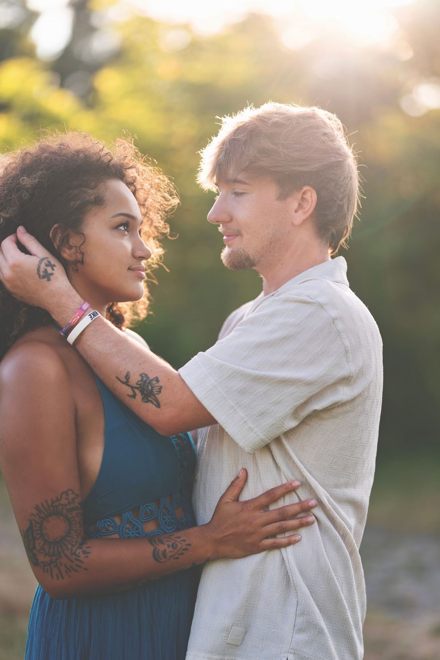 A romantic couple embraces outdoors during golden hour with soft sunlight filtering through trees in the background.