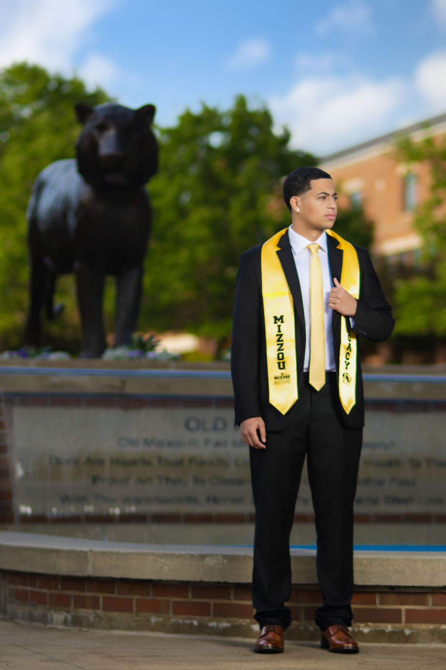Graduate in black suit with gold MIZZOU stole stands before bronze panther statue at University of Missouri.