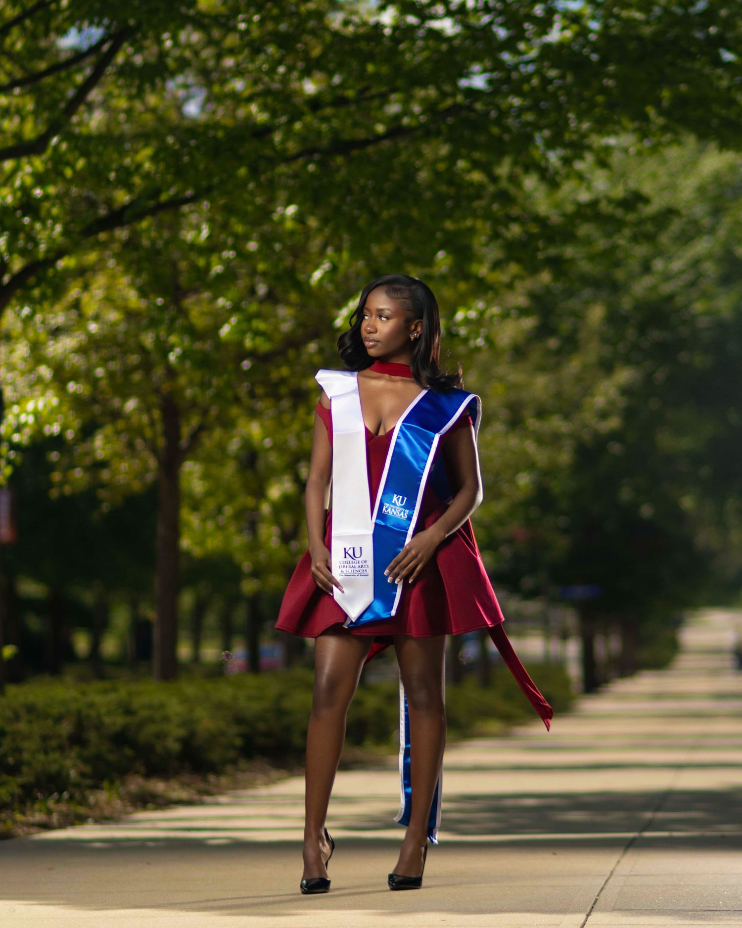 Young woman in maroon graduation dress and blue sash poses confidently on tree-lined campus walkway.