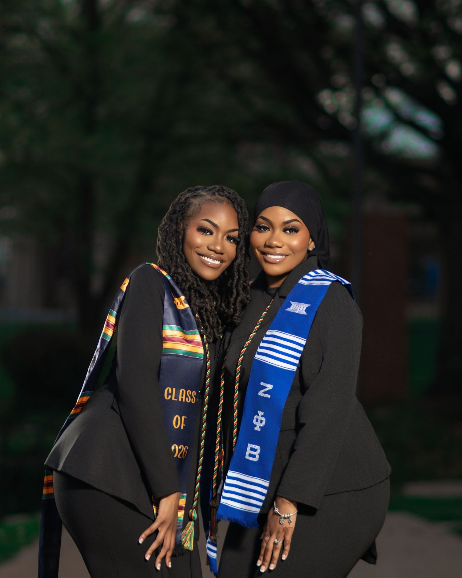 Two smiling graduates in black robes with colorful kente graduation stoles posing together.