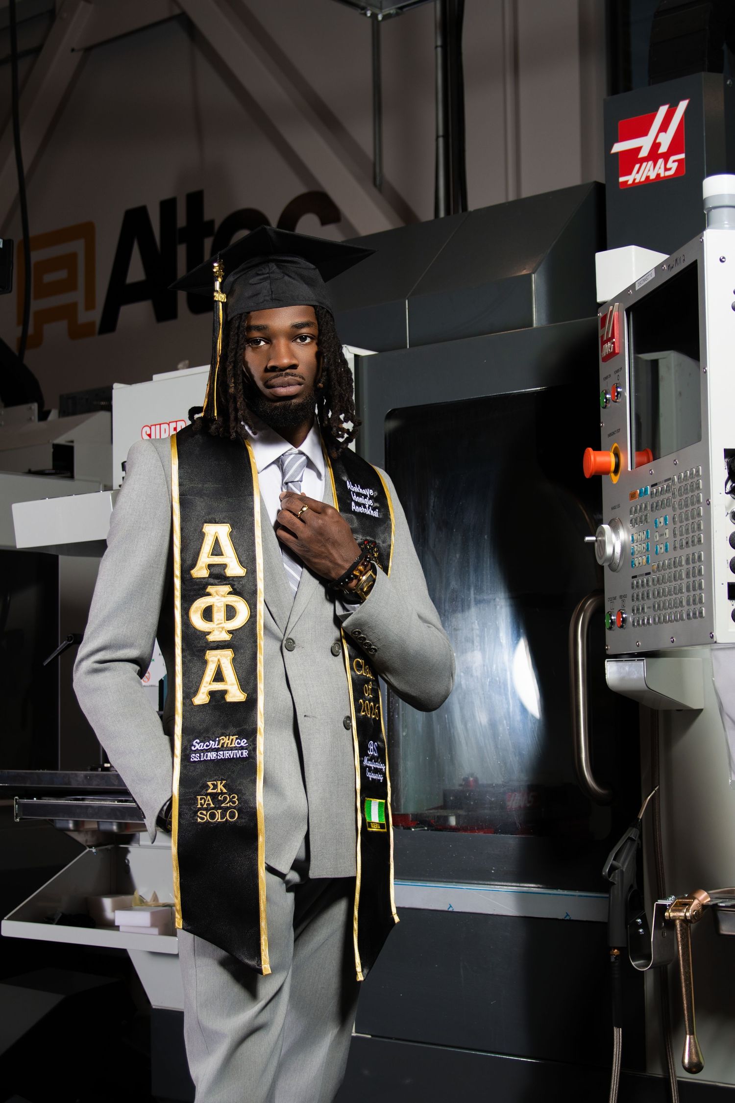 Graduate in cap and gown wearing Alpha Phi Alpha stole poses proudly in front of industrial Haas CNC machinery.