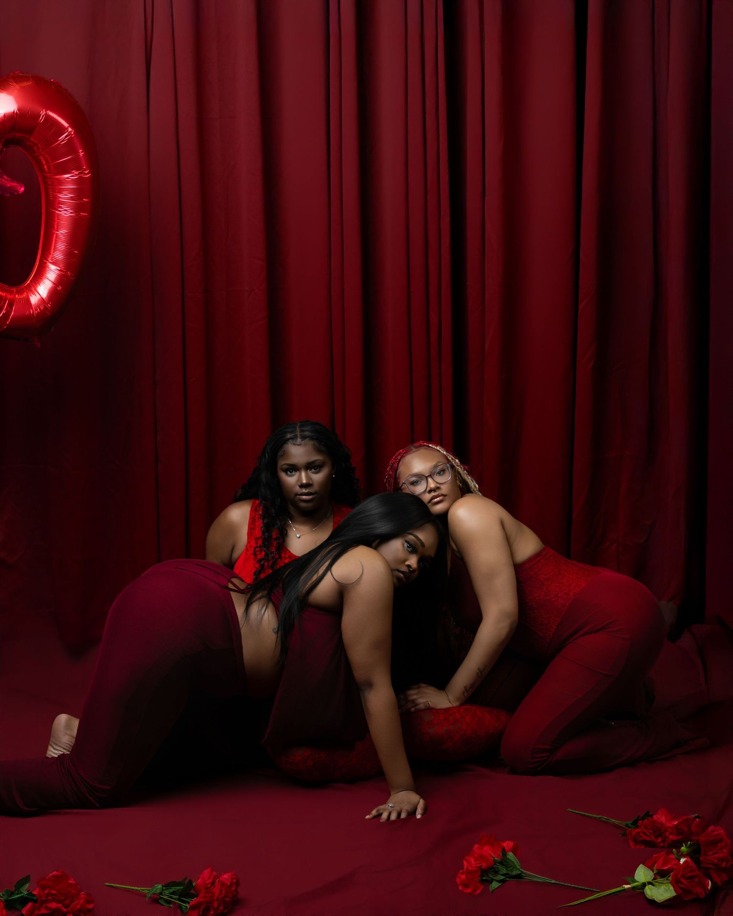 Three women in red outfits pose dramatically on floor with roses and heart balloon against red curtain backdrop.